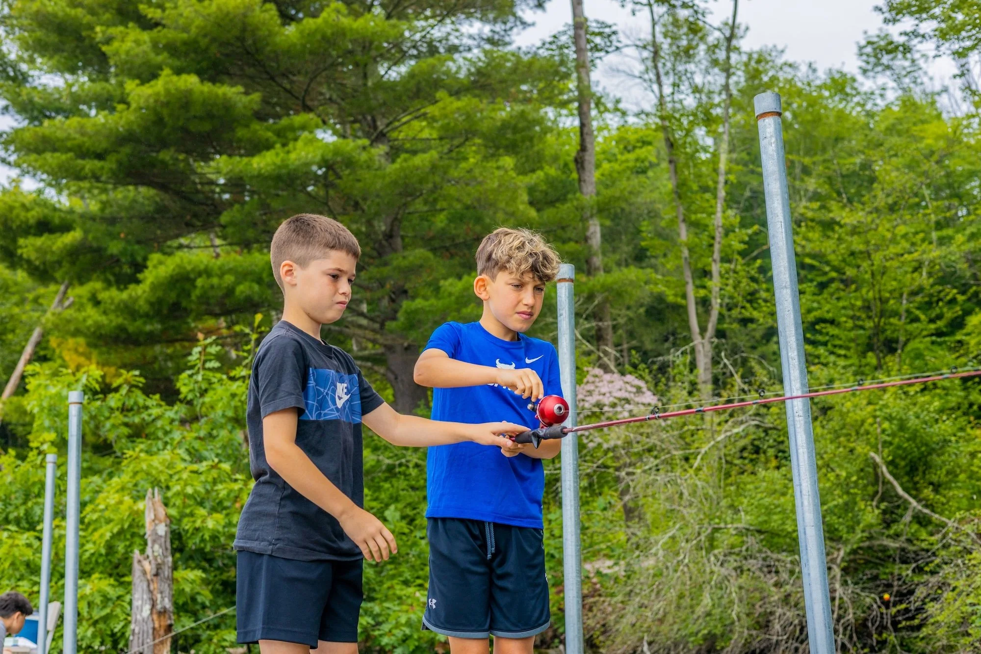 Two boys fishing with a red fishing pole outdoors surrounded by green trees.