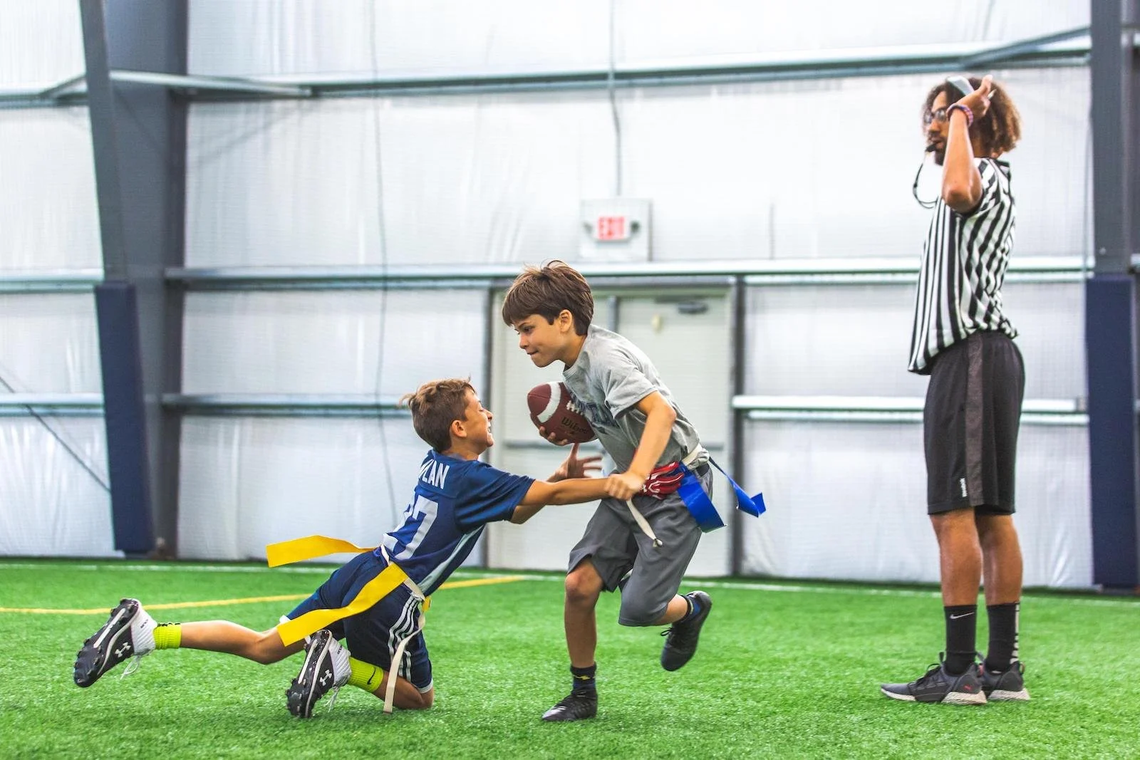 Two young boys playing flag football indoors, one running with a football while the other attempts to pull his flag, with a referee watching in the background.