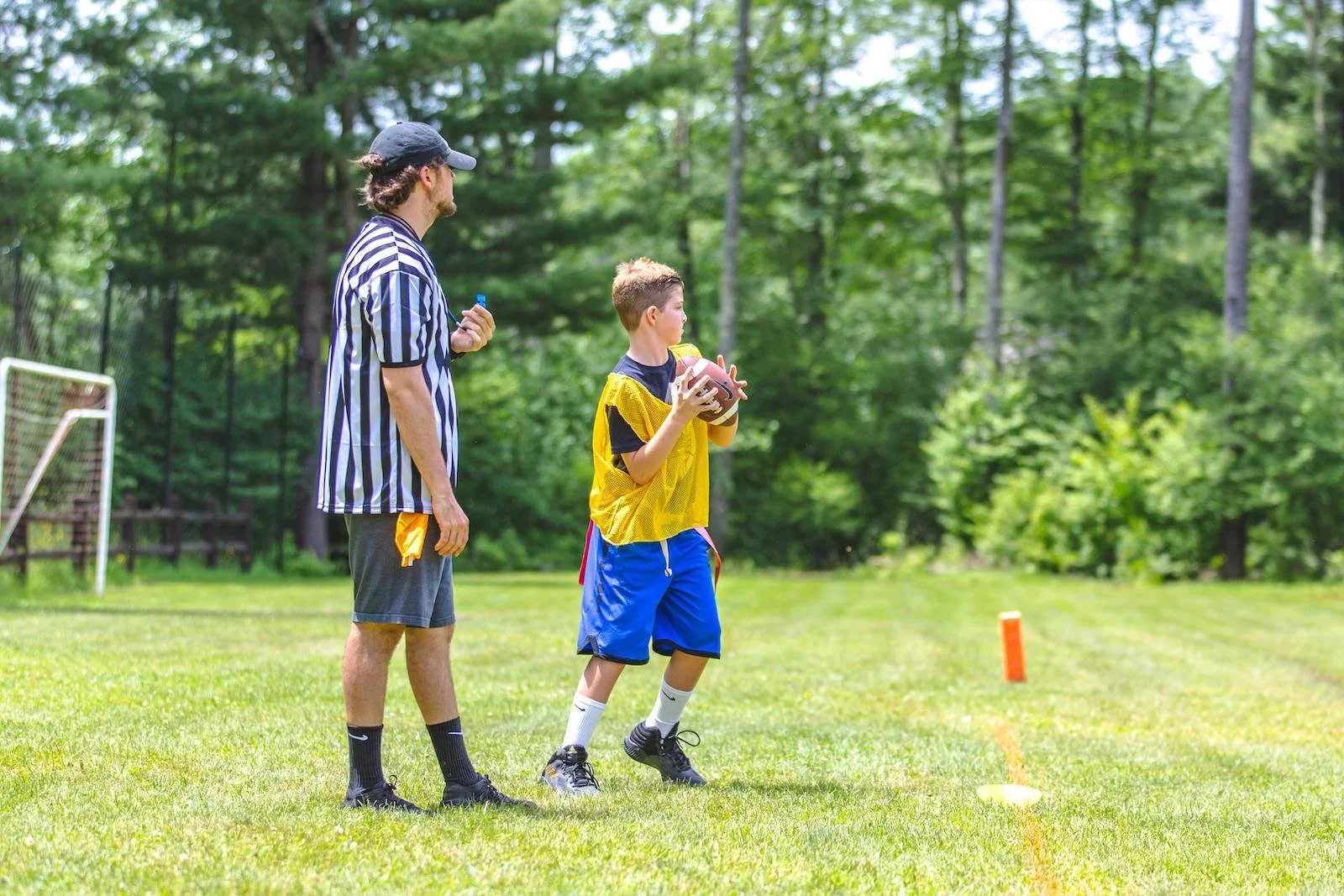 A young boy in a yellow jersey preparing to catch a football with a coach or referee nearby, on a grassy field with trees in the background.