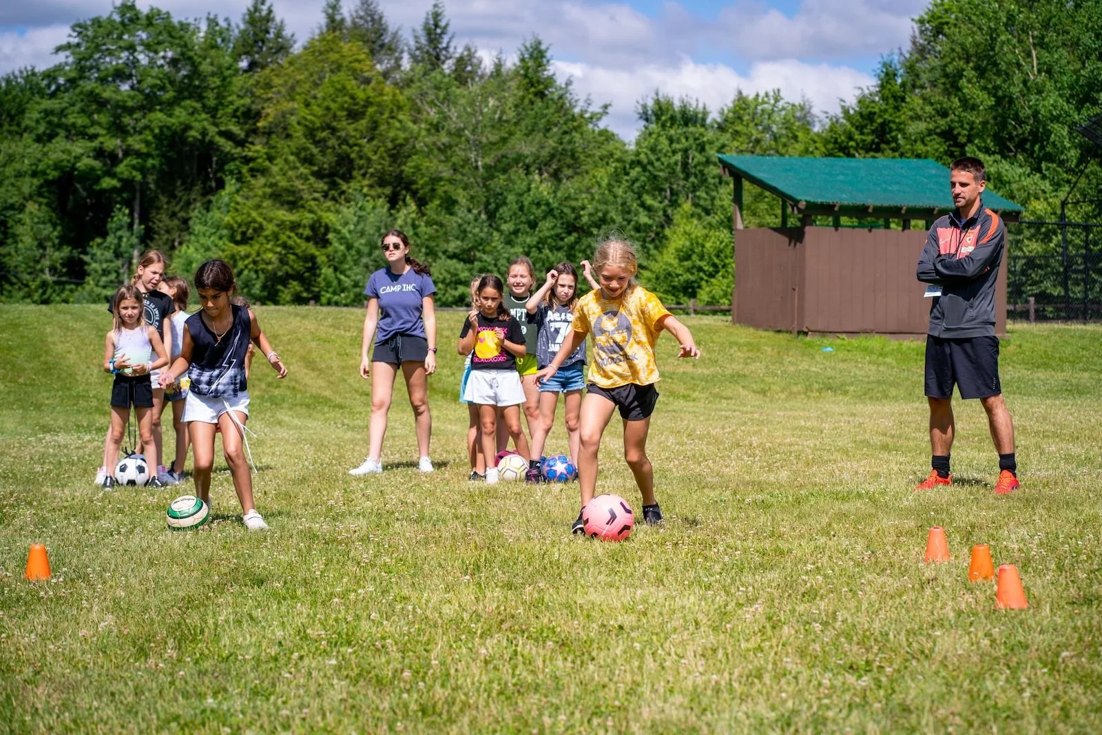 A group of young girls participating in a soccer drill on a grassy field, with a coach supervising, surrounded by trees and a blue sky.