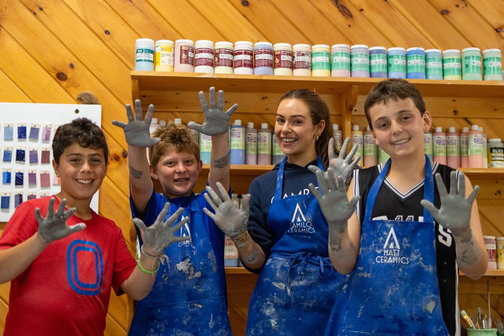 Four children and a young woman smiling and showing their hands covered in gray ceramic clay in a ceramics painting studio. They are wearing blue aprons with 'Matt Camilo Ceramics' logos, and behind them are shelves with colorful ceramic paint bottle