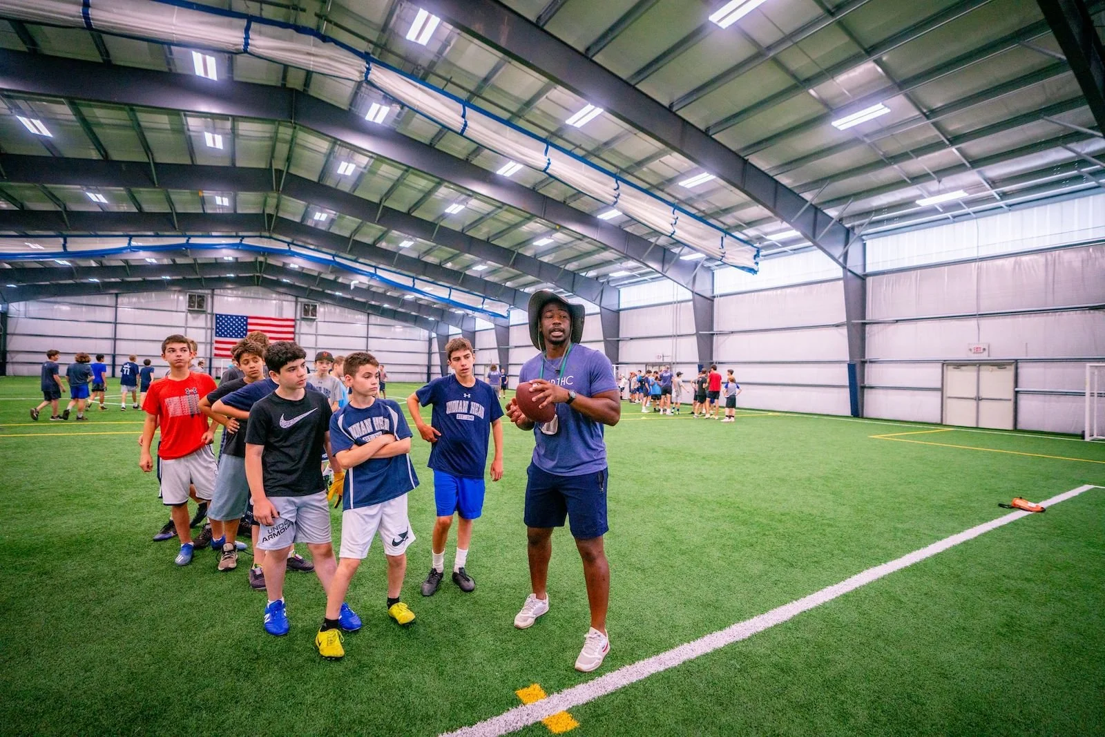 Football coach in a hat giving instructions to a group of young athletes in a spacious indoor turf football field.