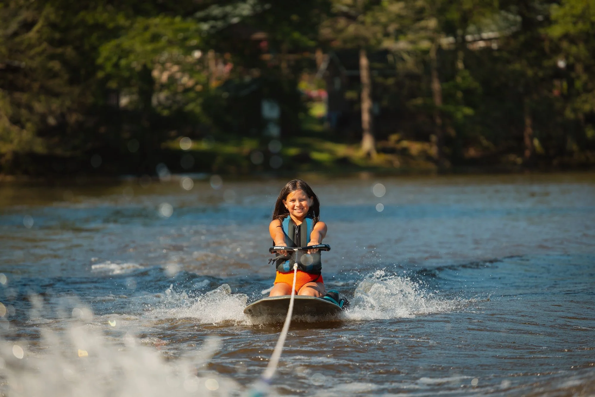 A young girl in a life jacket riding a wakeboard on a lake with trees in the background, smiling and holding onto a tow rope.