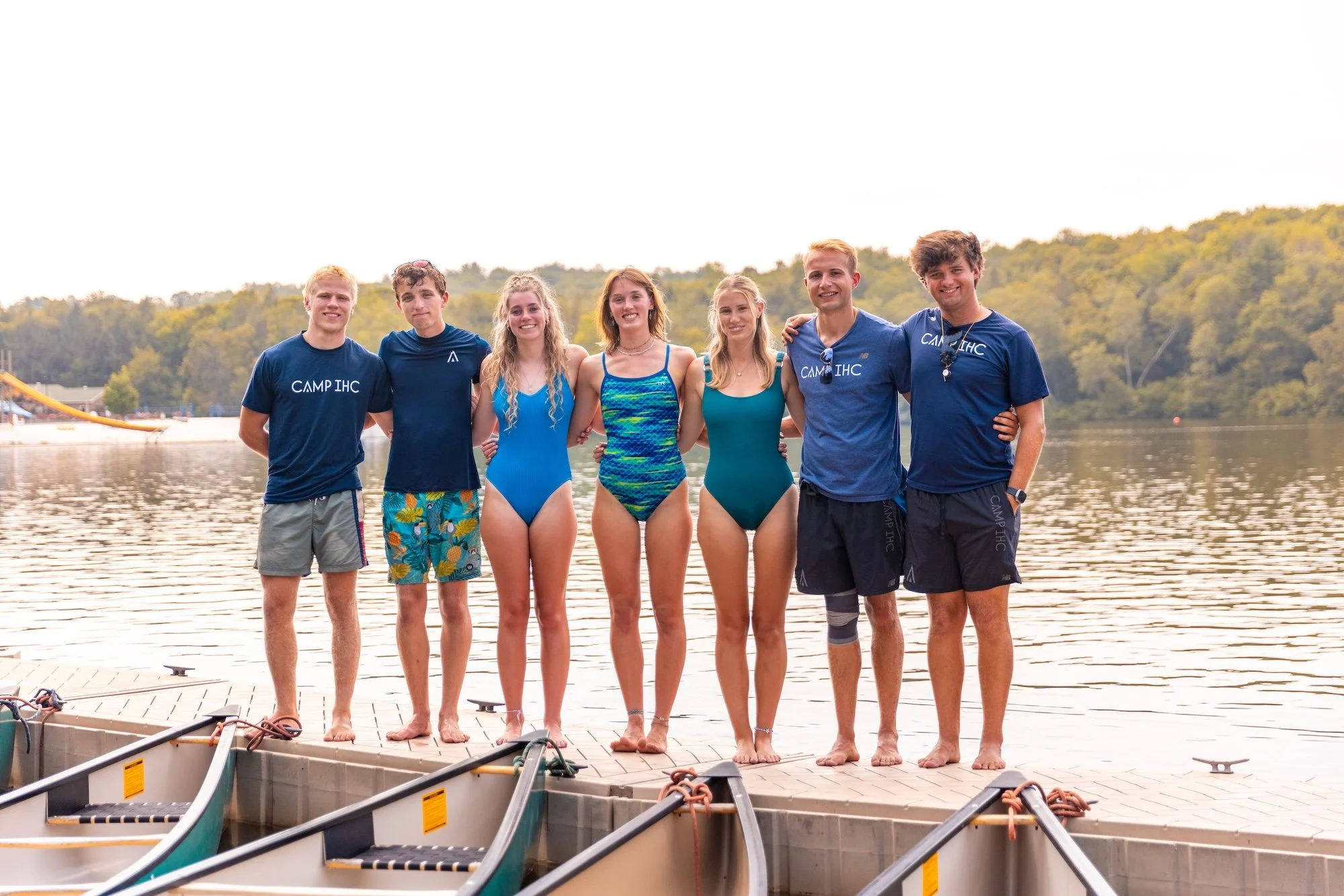 A group of seven young people standing hand-in-hand on a dock near a lake, smiling at the camera, with canoes in the foreground and trees in the background.