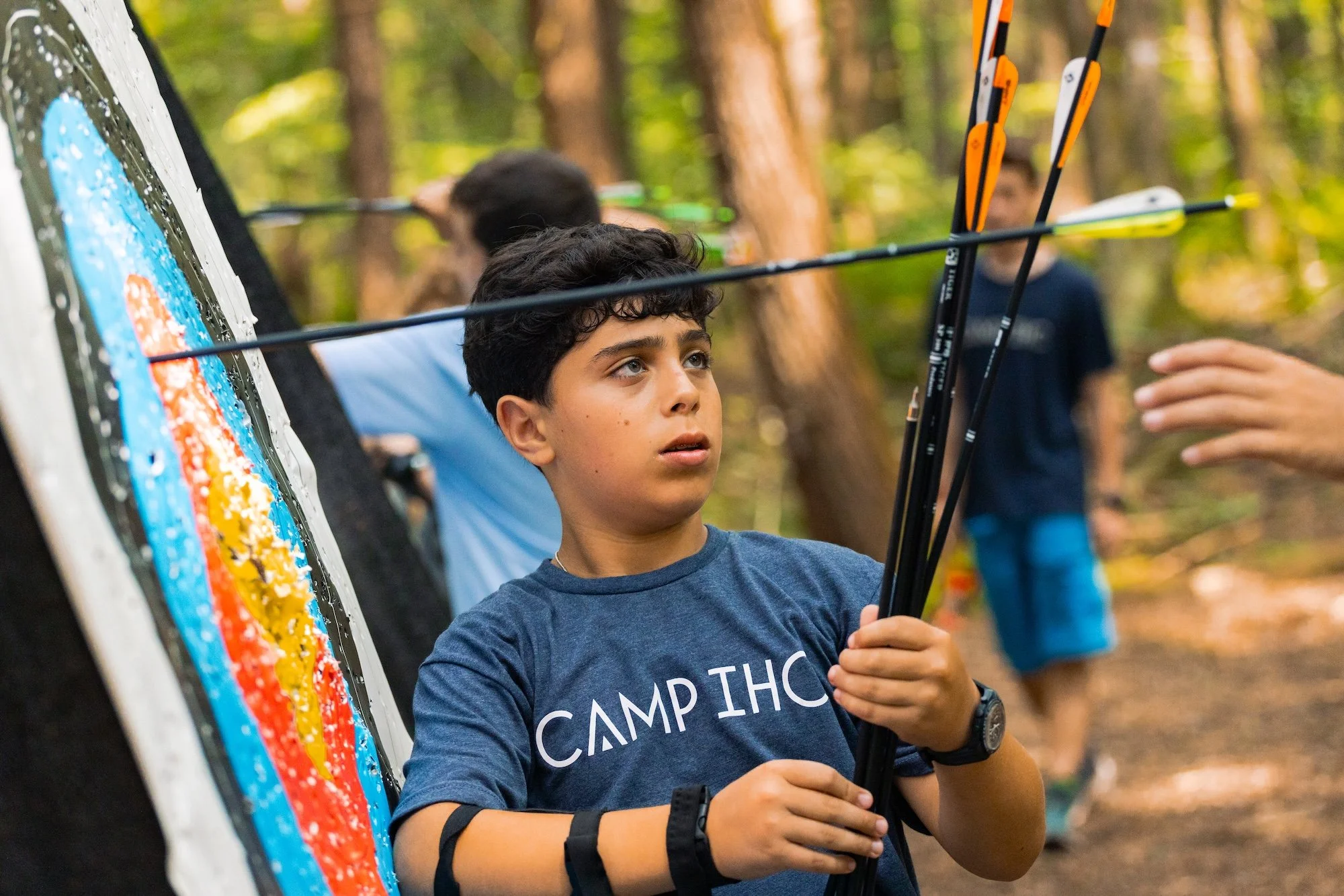 A boy at a camp shooting arrows at a target with other children in the background in a wooded area.