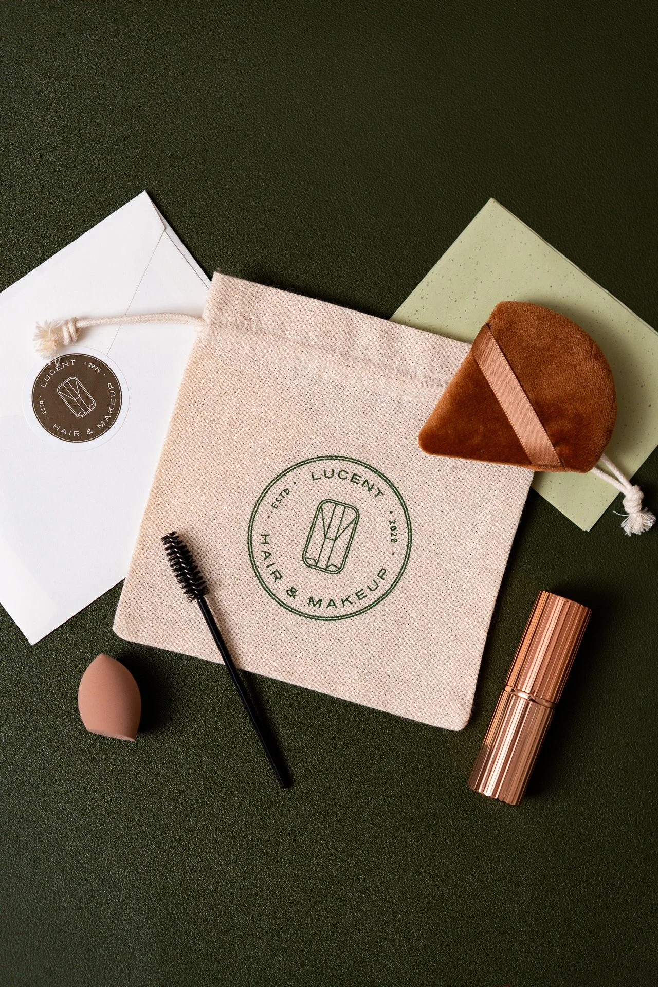 Beauty and makeup products including a makeup sponge, mascara wand, an eyelash curler, a lipstick, and branded packaging with a drawstring bag and envelopes on a dark green surface.