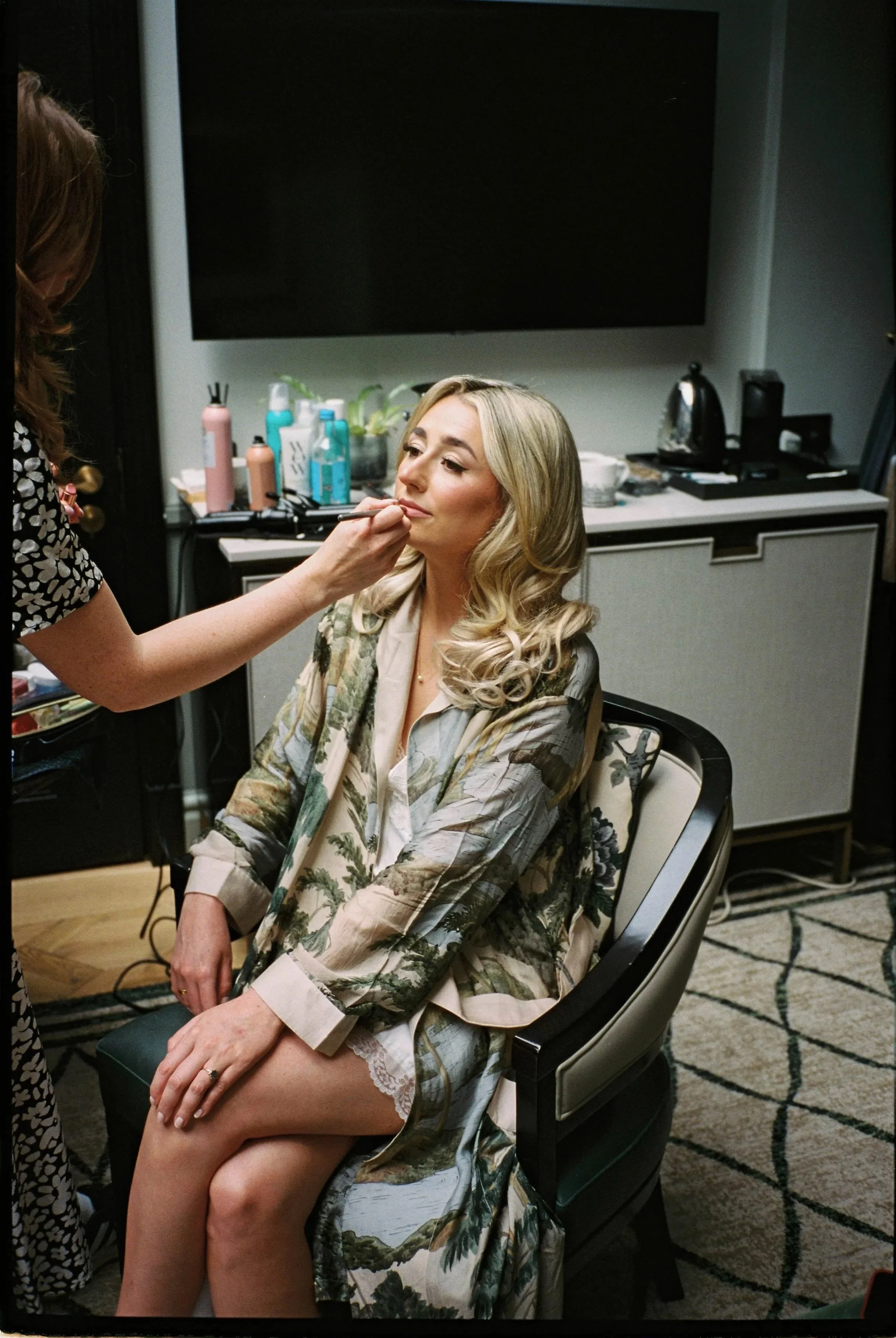 A bride with blonde hair sitting in a chair, having her makeup done by a makeup artist in a London hotel room