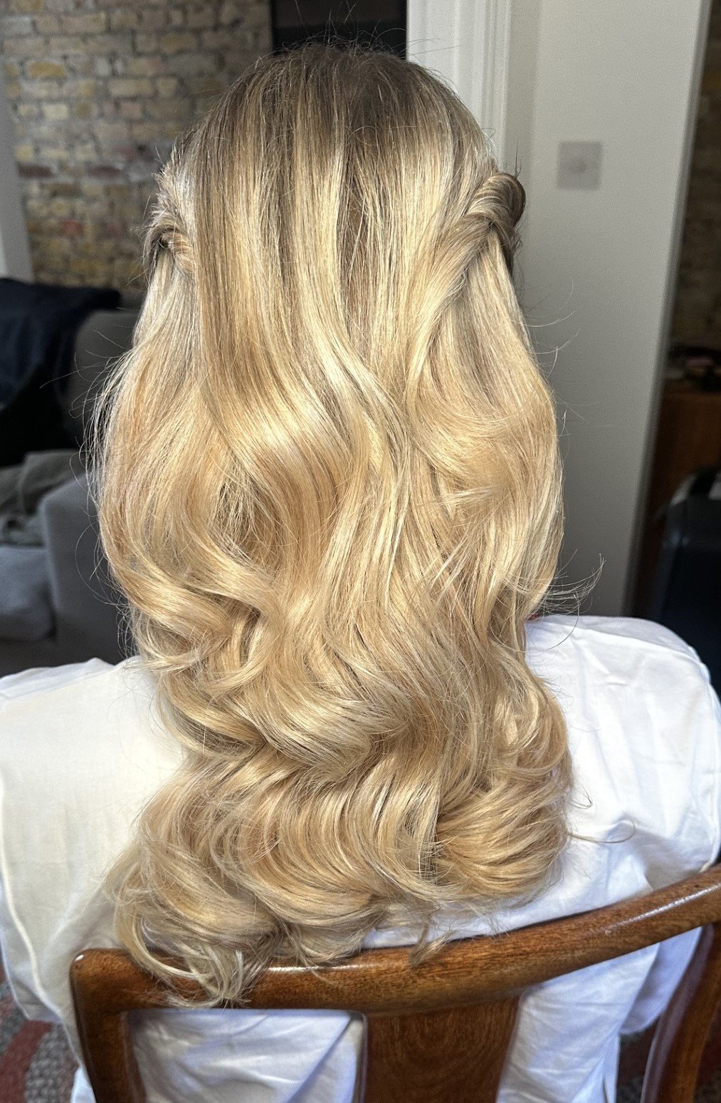 Back view of a woman with long, wavy blonde hair, styled with soft waves, sitting on a wooden chair in a room with exposed brick wall.