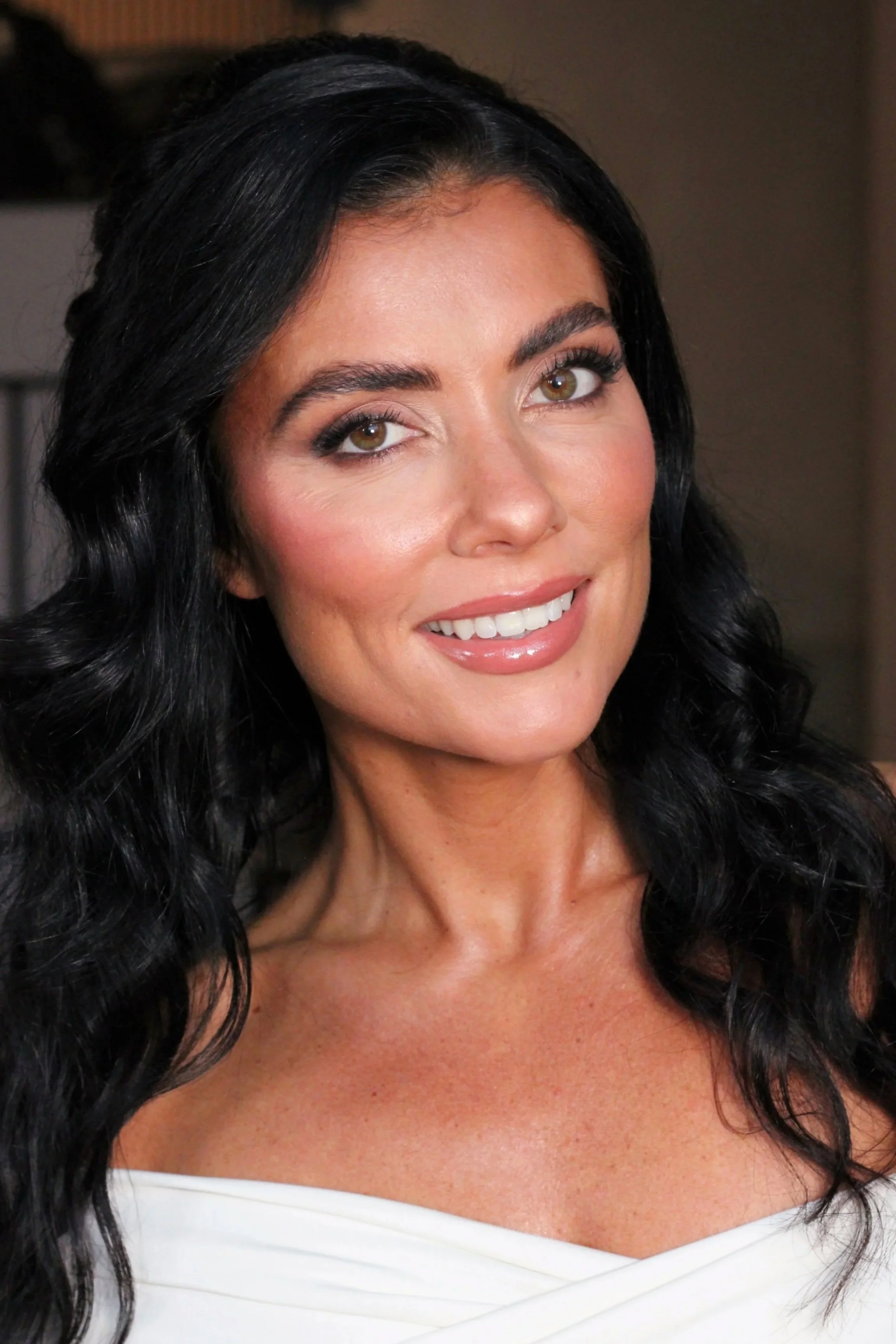 A woman with long, wavy black hair smiling at the camera, wearing soft glam bridal makeup.