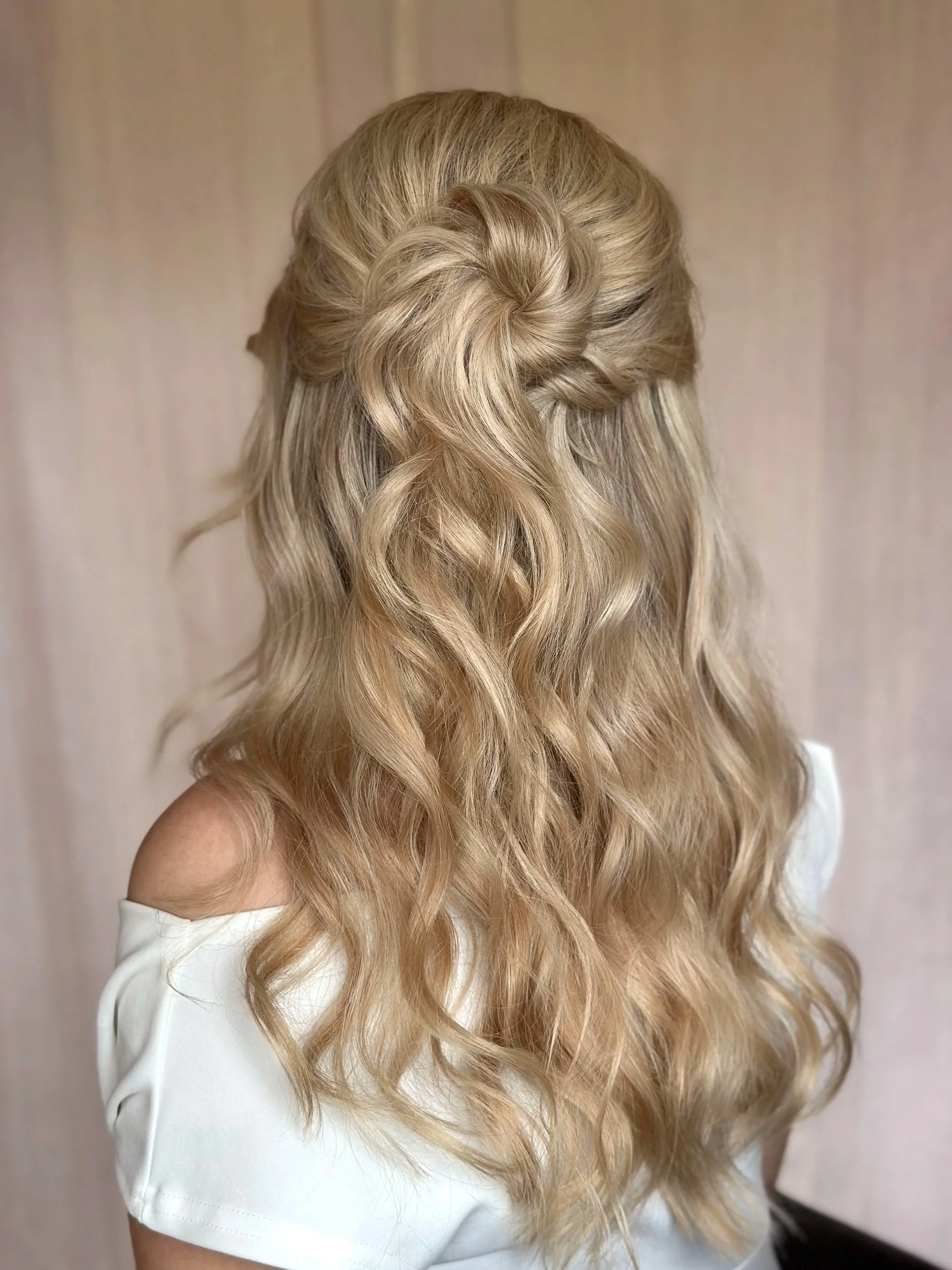 Back of woman's head with styled blonde wavy hair, featuring a twisted knot and natural waves, wearing a white off-the-shoulder top.