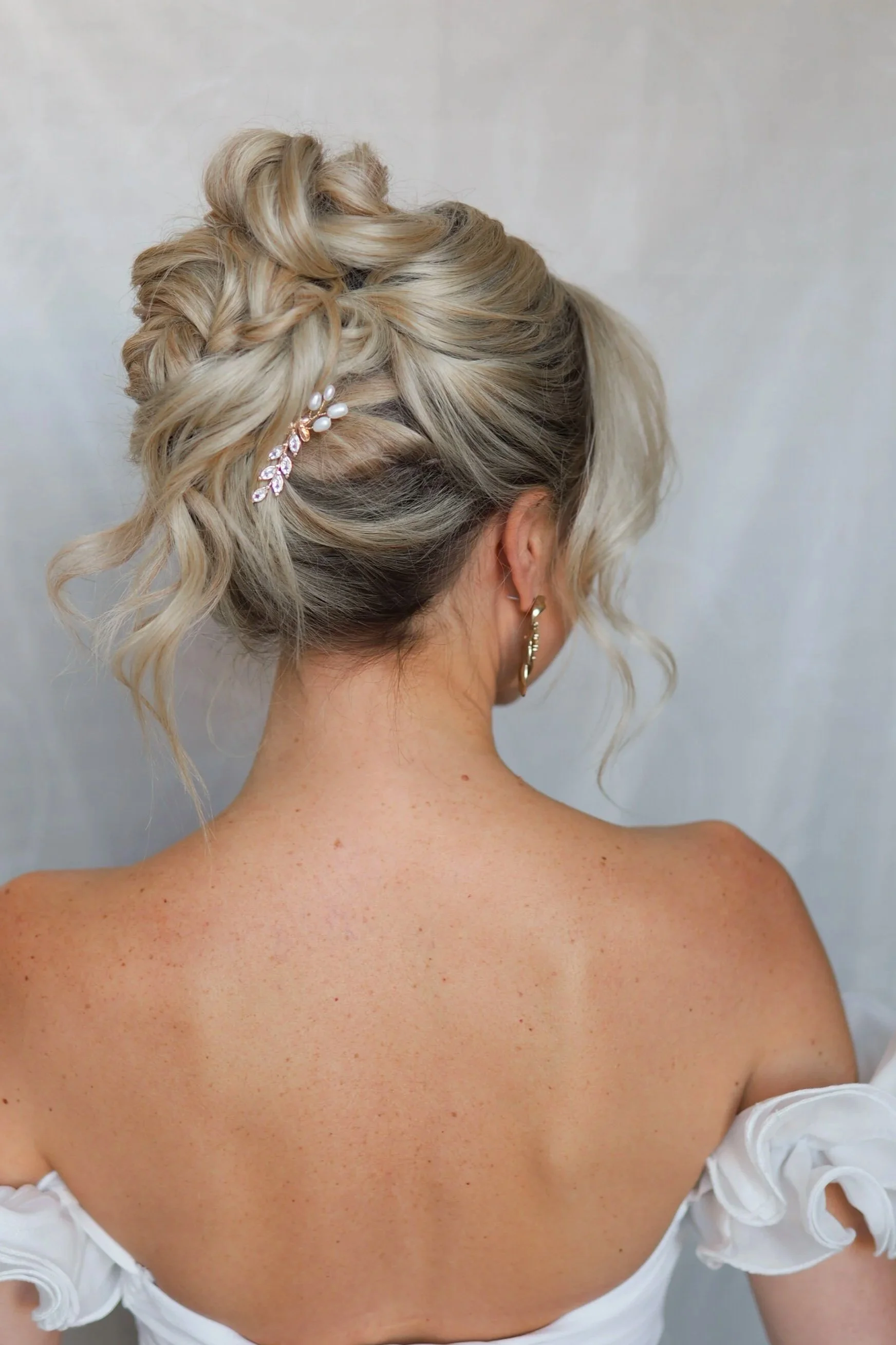 Back view of a woman with blonde, boho updo hairstyle decorated with a pearl and rhinestone hairpin, wearing a white off-shoulder dress and gold hoop earrings.