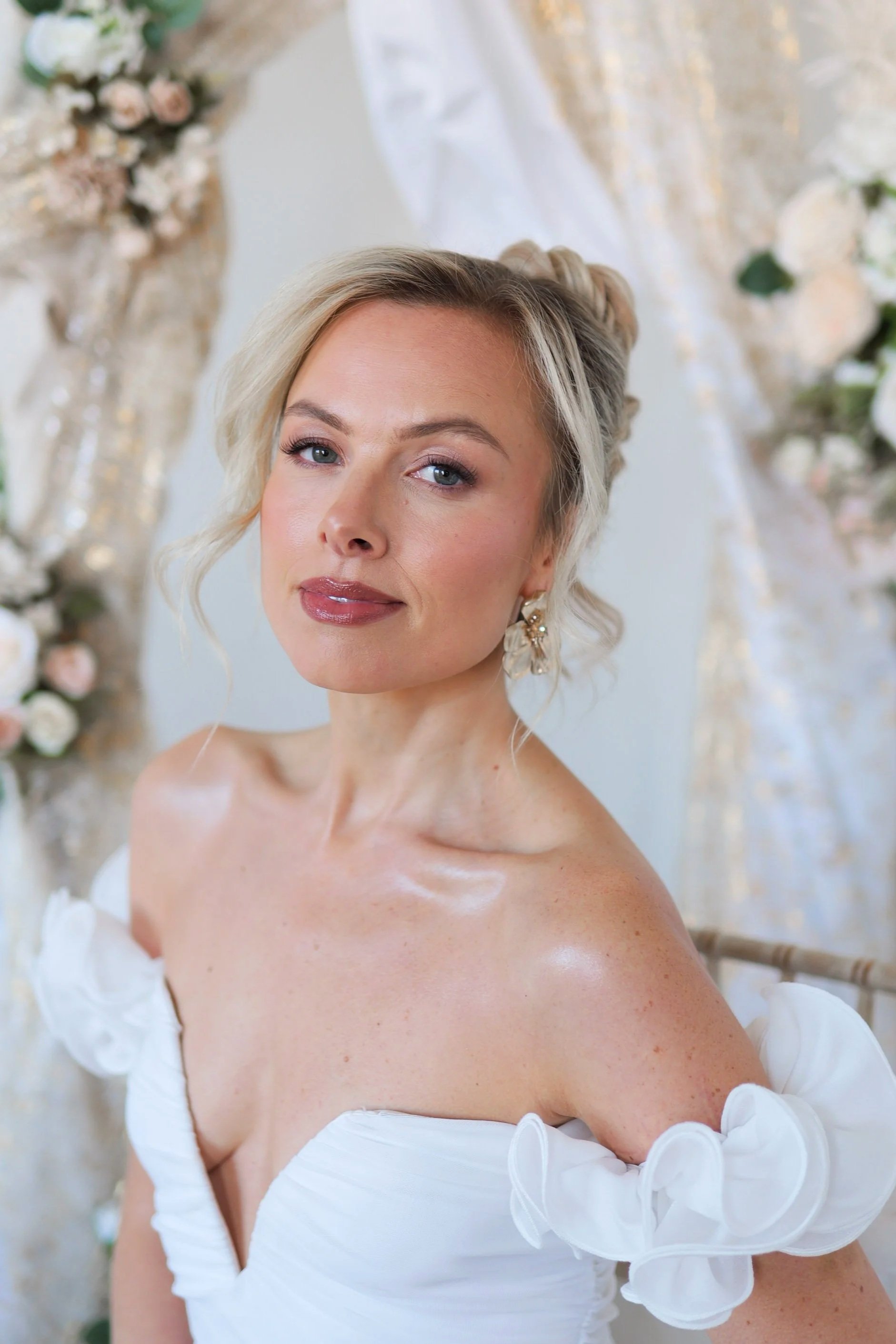 A woman with blonde hair styled in waves, wearing a white off-the-shoulder dress with ruffled sleeves and earrings, posing in front of a floral backdrop.