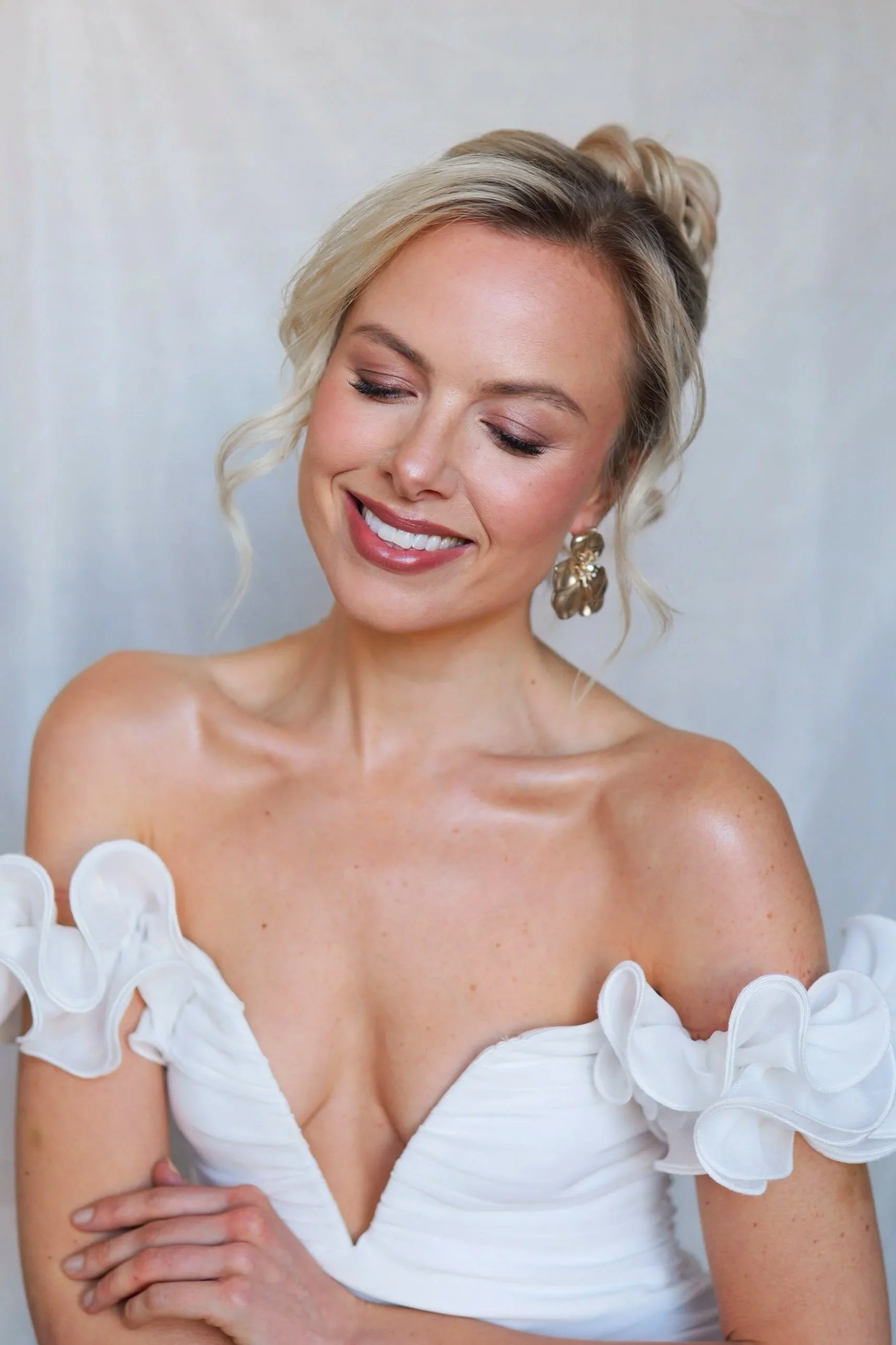 A smiling bride with blonde hair styled in an updo, wearing large gold earrings and an off-shoulder white dress with ruffled sleeves, posing against a plain background.