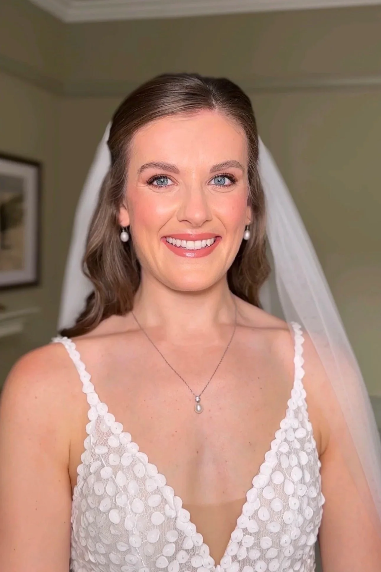An image of a bride smiling at the camera on her wedding day. She wears a white wedding dress and veil, with soft glam makeup and an elegant half-up hairstyle. 