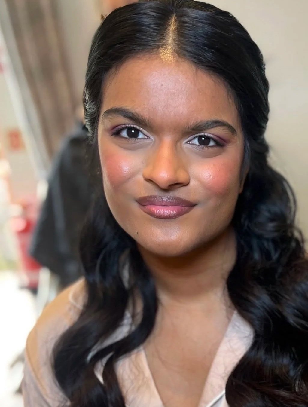 Close-up portrait of a woman with long, dark, wavy hair and a confident expression, wearing natural makeup, in an indoor setting.