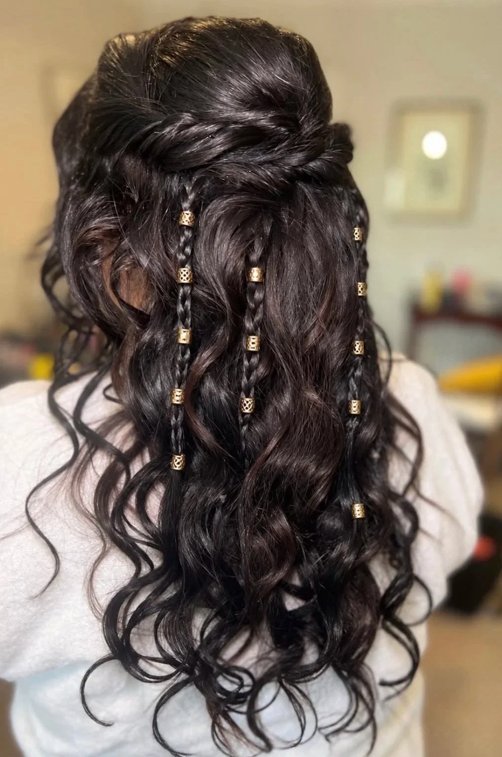Back view of a woman with long, dark, wavy hair decorated with multiple small gold hair cuffs, some of which have a lattice design, creating a braided and wavy boho hairstyle.