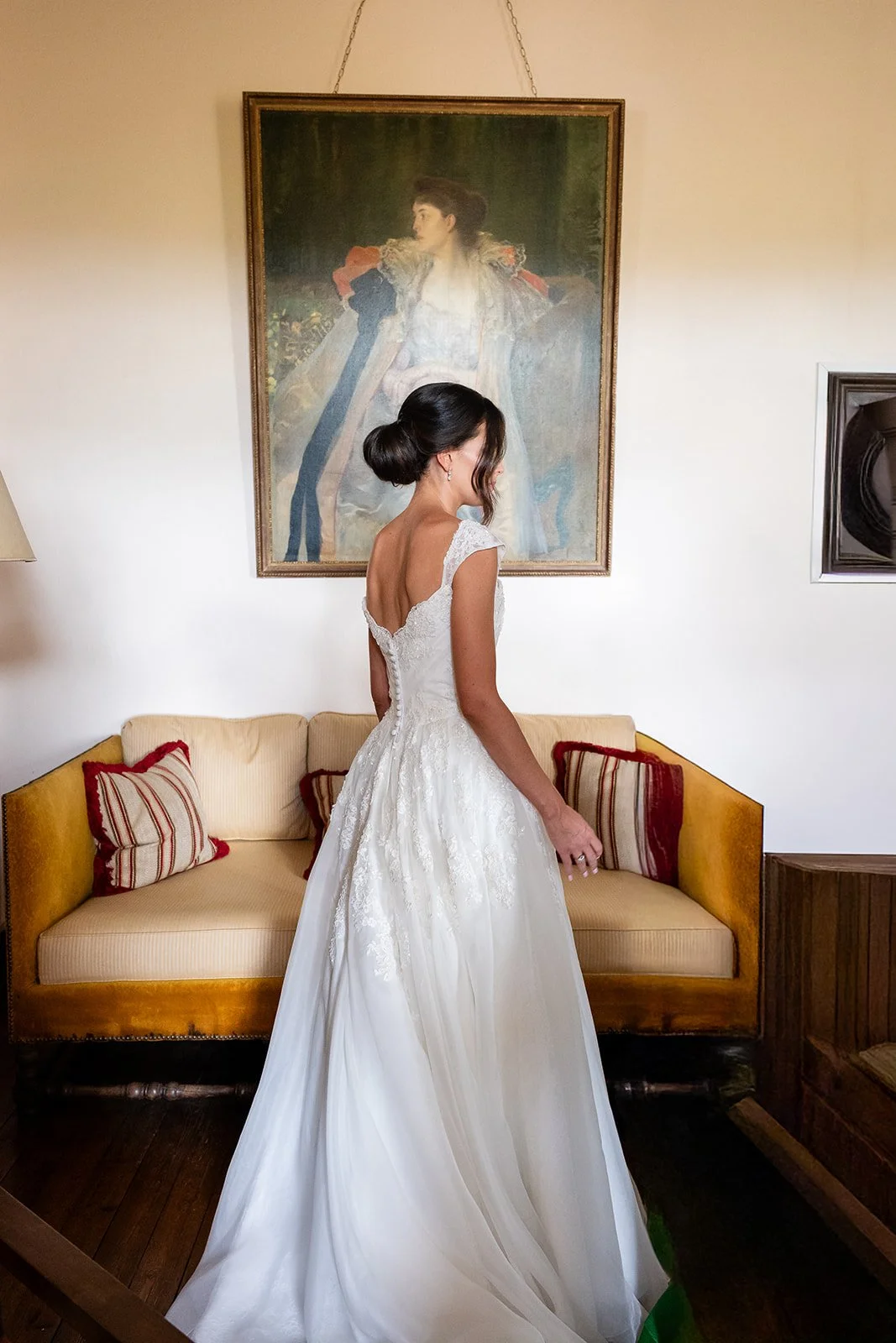 A bride stands in front of a sofa, with a painting on the wall behind her. She wears a white wedding dress and has her hair styled in an elegant, low updo.