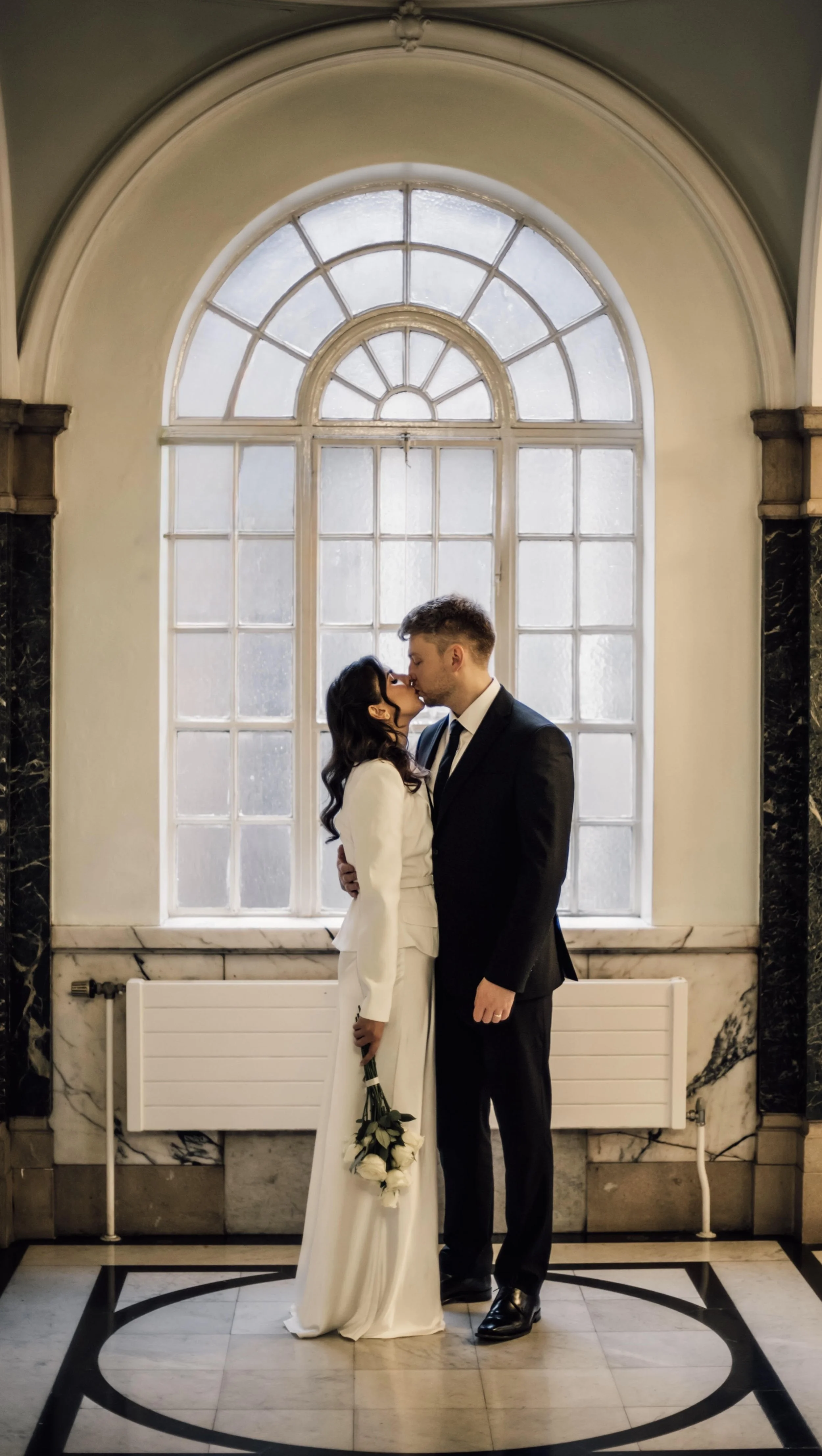A newlywed couple sharing a kiss inside a London wedding venue, standing in front of a large arched window with frosted glass, surrounded by marble and stone exterior, with a black and white tiled floor.