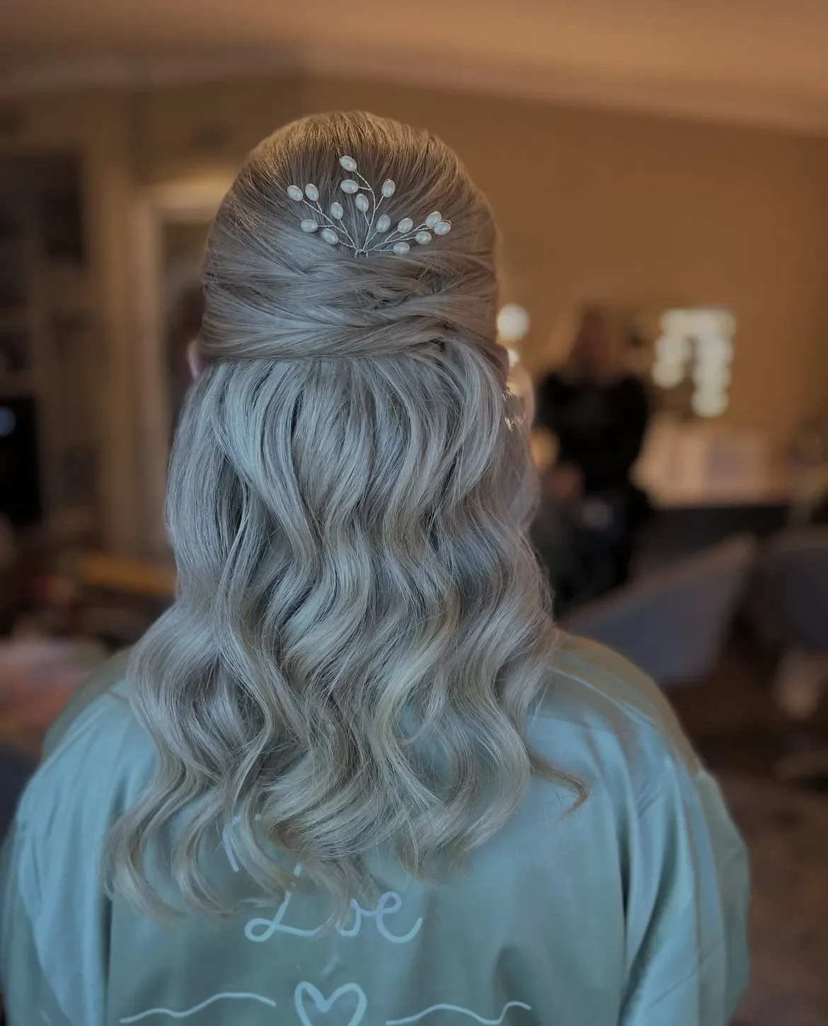 Back of a person's head with long, wavy blonde hair, styled in a half-up with twists, decorated with a pearl hairpin, in a warmly lit indoor setting.