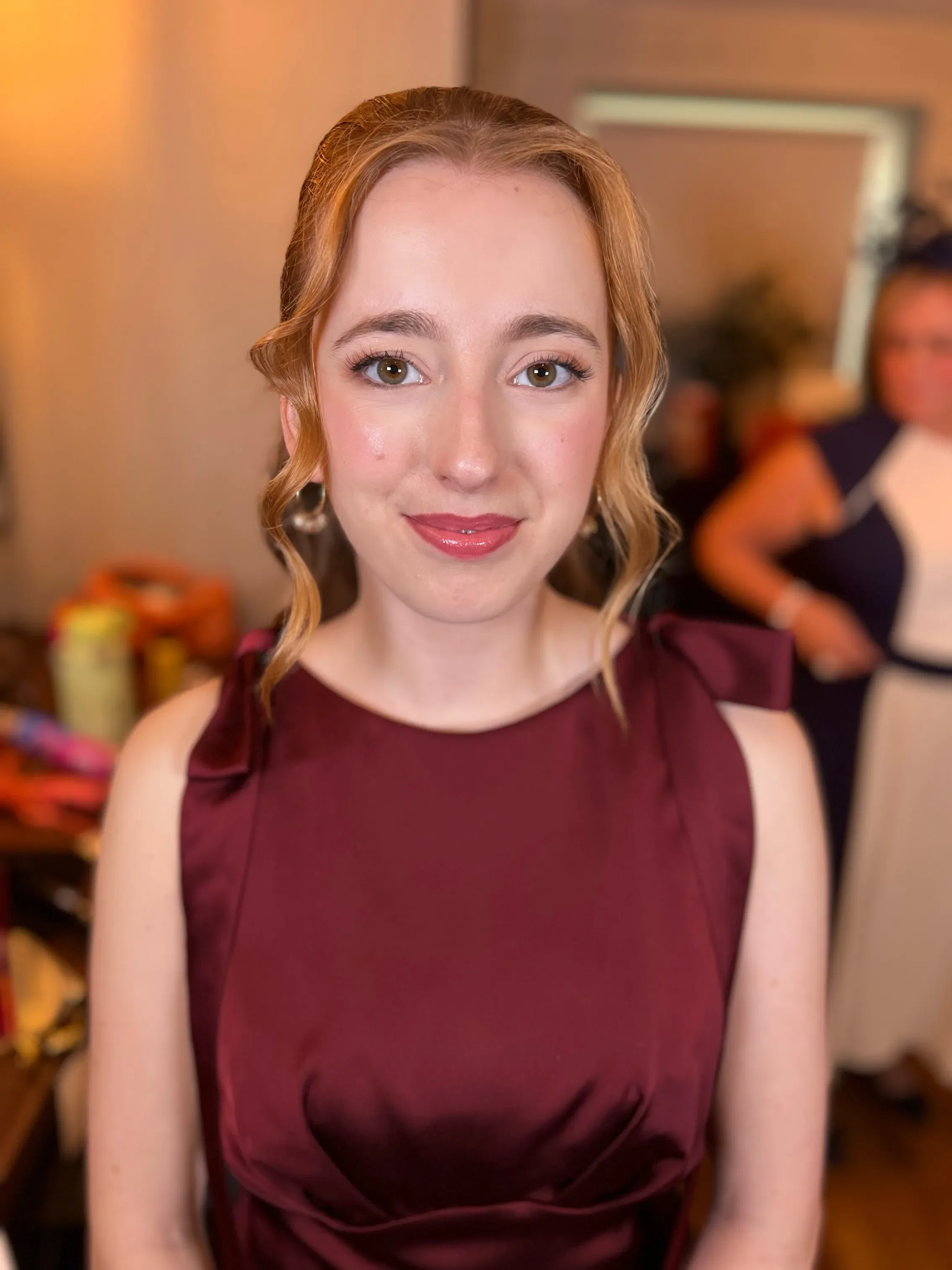 Close-up of a young bridesmaid with light skin, brown eyes, and curly red hair wearing a maroon dress, smiling indoors with a blurred background.