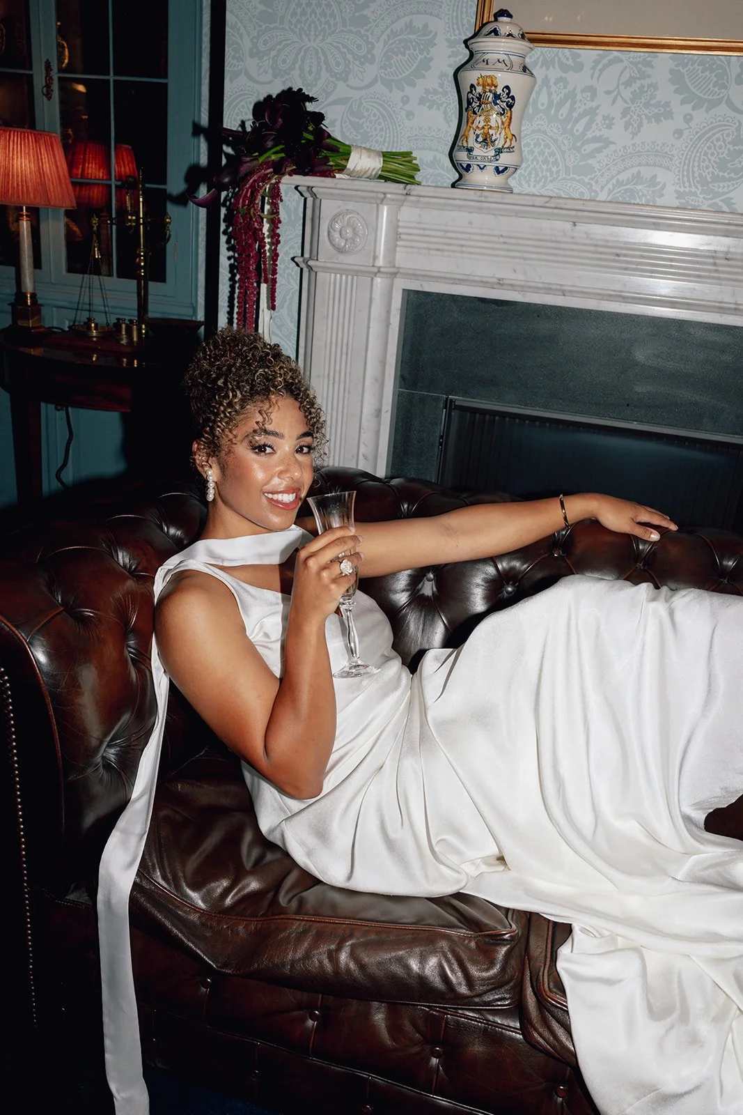 A woman in a white wedding gown, sitting on a leather couch, holding a champagne glass, smiling.