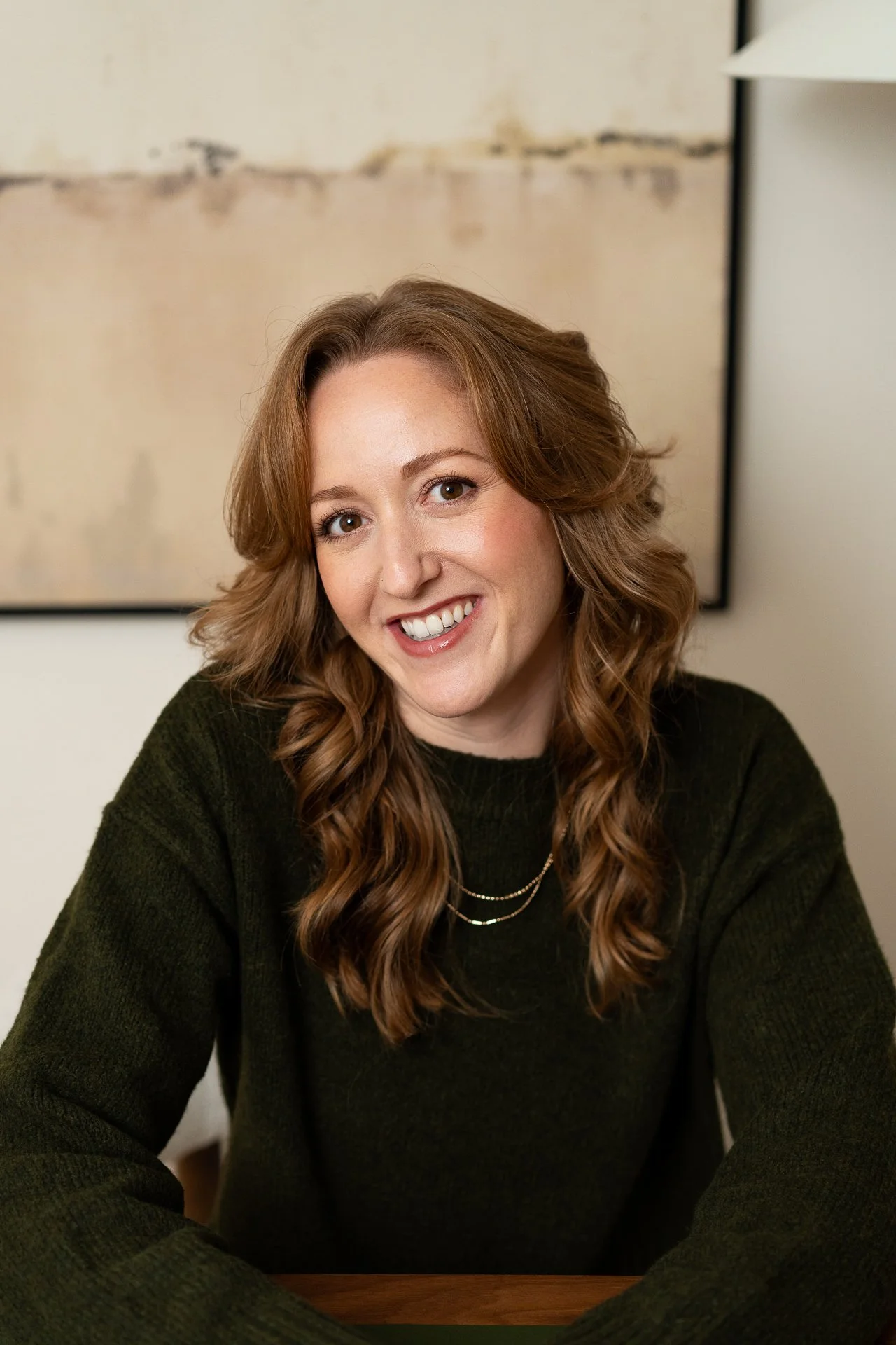 A woman with shoulder-length wavy auburn hair, smiling while sitting at a table indoors, wearing a dark green sweater and gold necklace.
