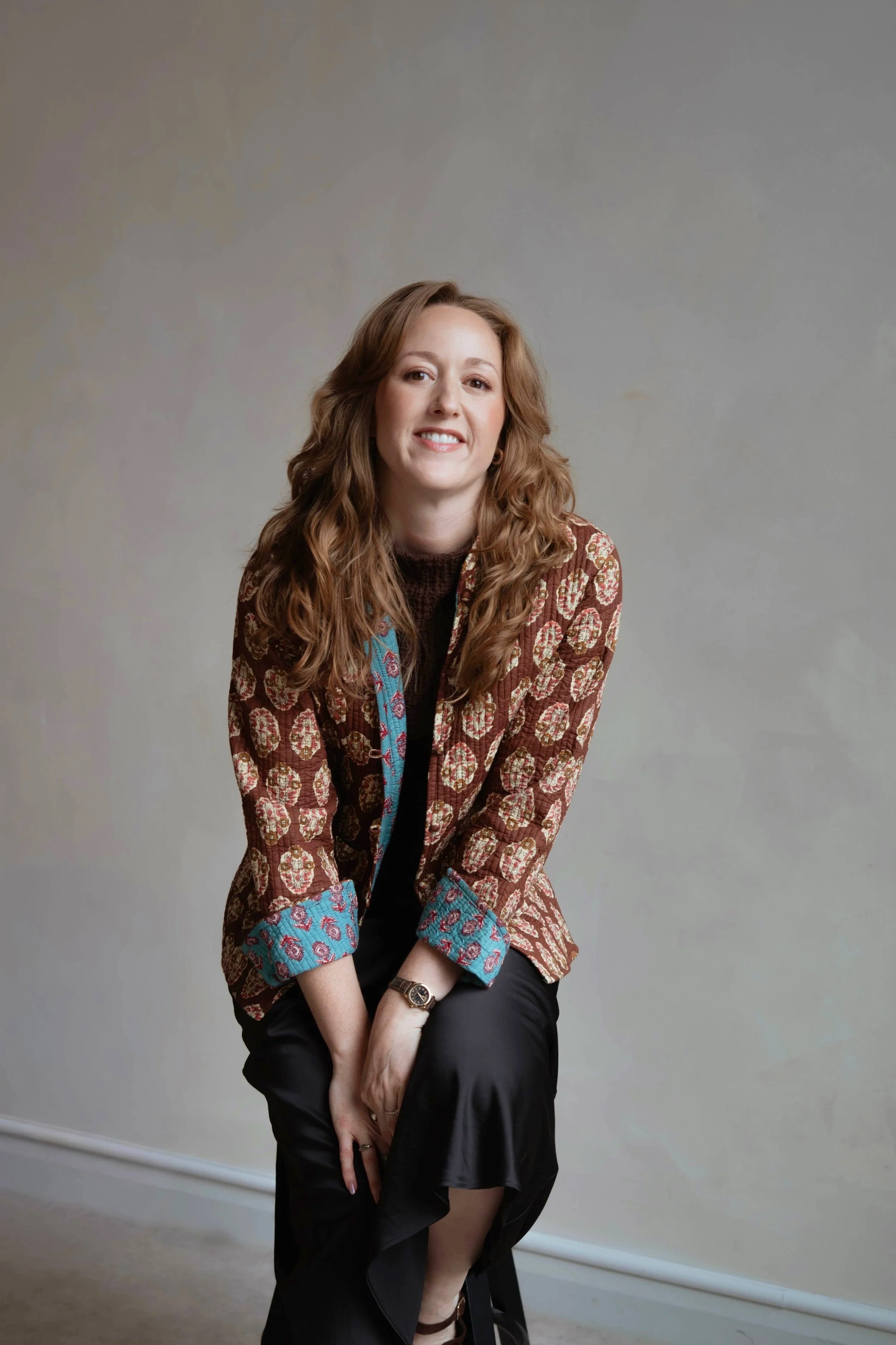 Image of a woman with auburn hair, sitting on a stool, wearing a patterned jacket and black skirt, smiling at the camera.
