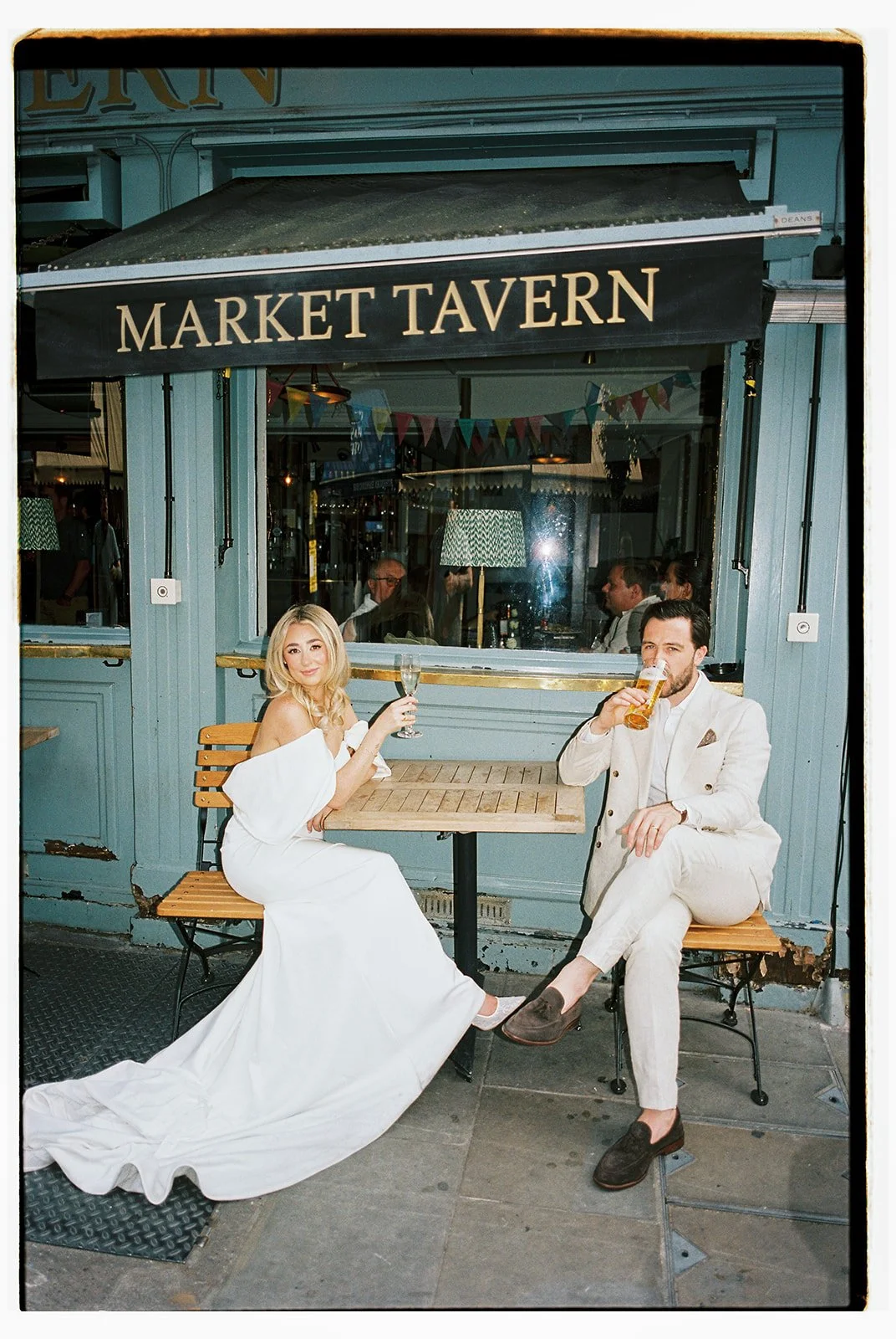 A woman in a white wedding dress and a man in a white suit sitting outside a bar labeled 'Market Tavern,' holding drinks and smiling.