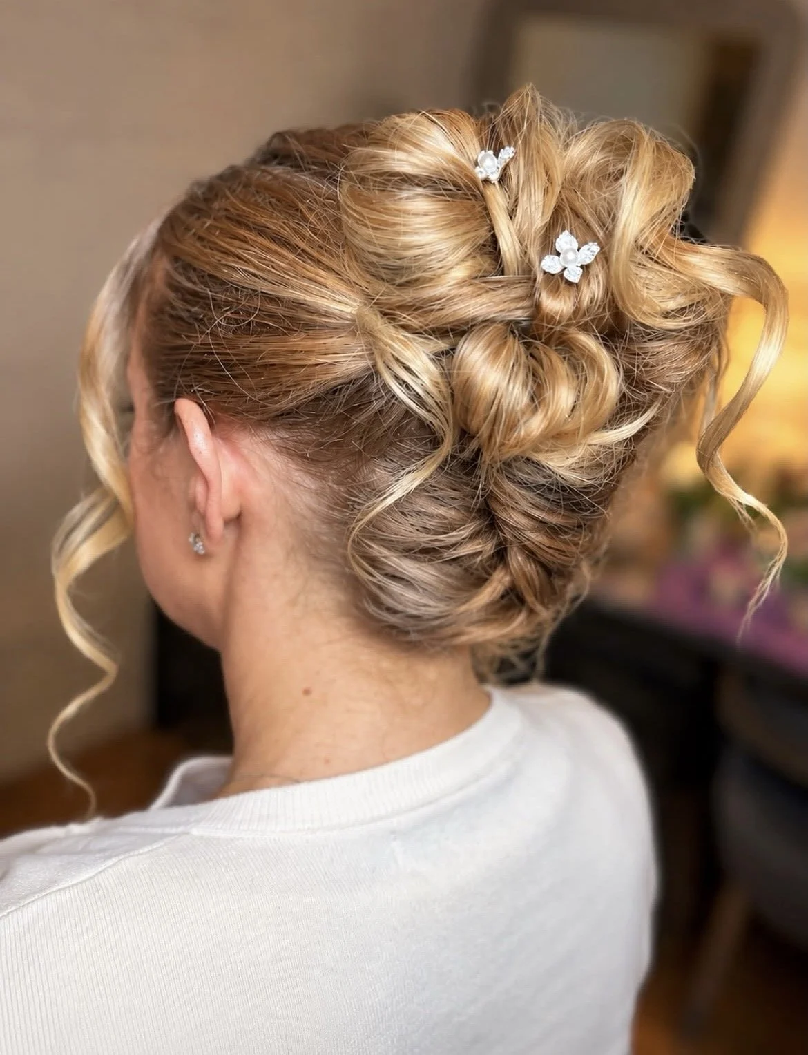 A woman with styled blonde hair in curls and updo, decorated with small white floral hair accessories, seen from behind, wearing earrings and a white top.