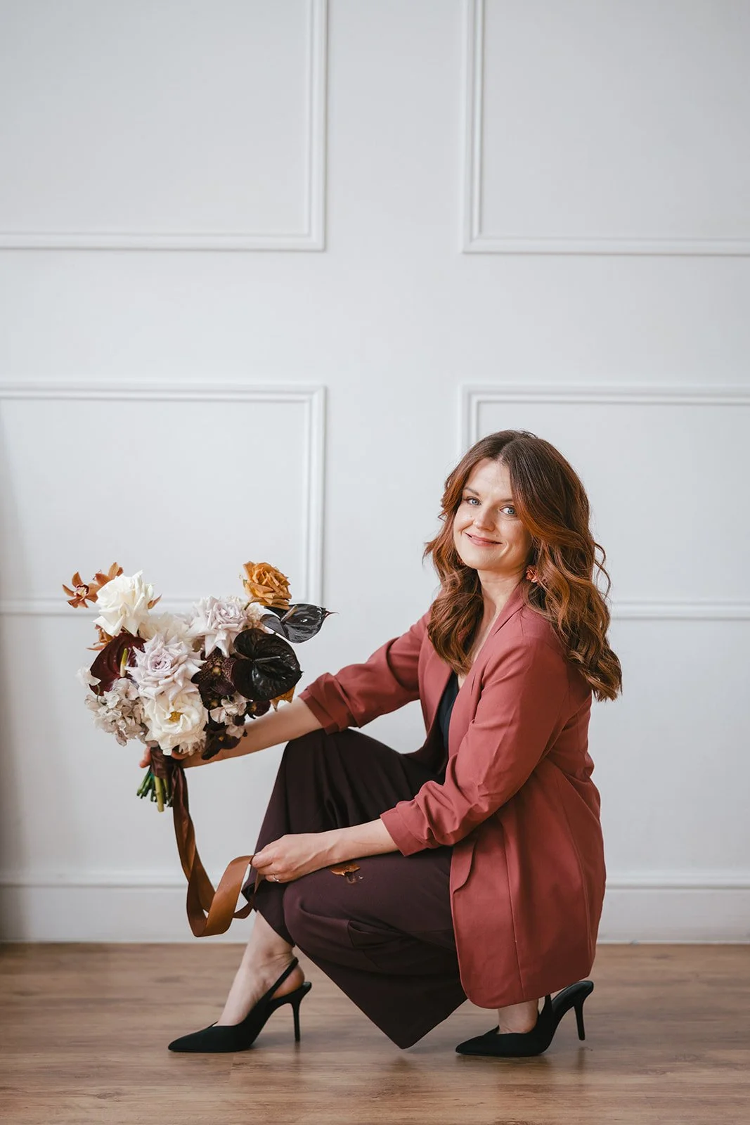 woman with red hair wearing a maroon blazer, black top, maroon trousers, and black high heels, sitting on the floor holding a bouquet of flowers, against a white panelled wall for a branding shoot