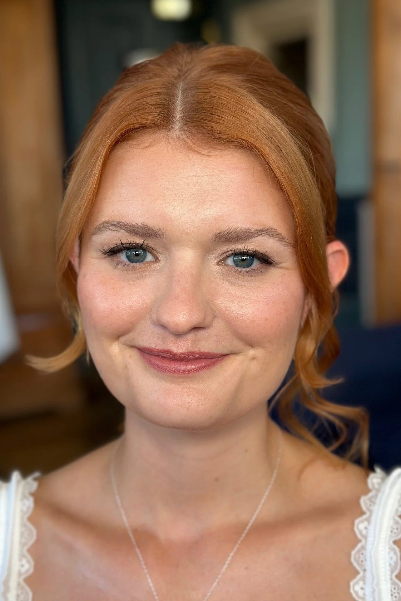 Close-up portrait of a woman with red hair, blue eyes, and a gentle smile, wearing a white lace-trimmed top and a delicate silver necklace.