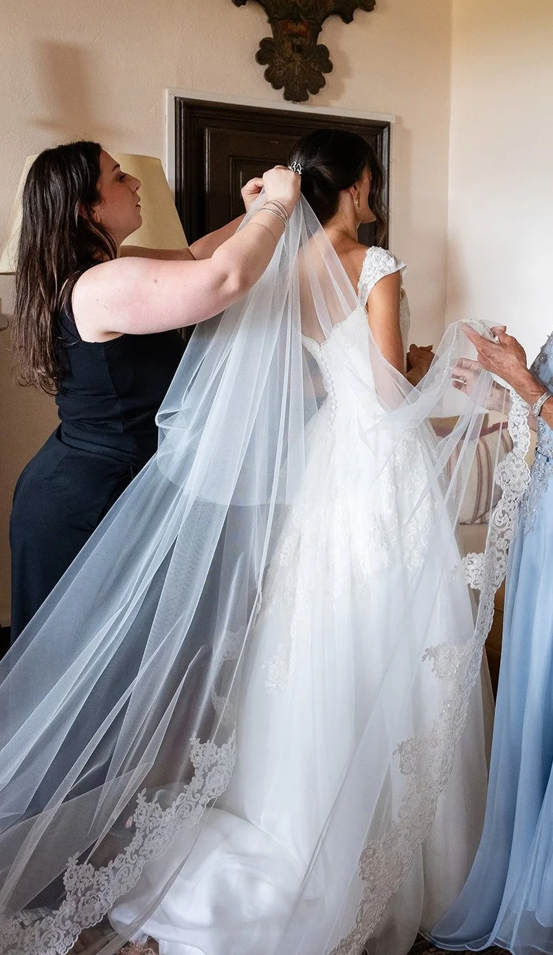 A hair and makeup artist is attaching a veil in a bride's hair on her wedding morning, the bride is wearing a white wedding dress 