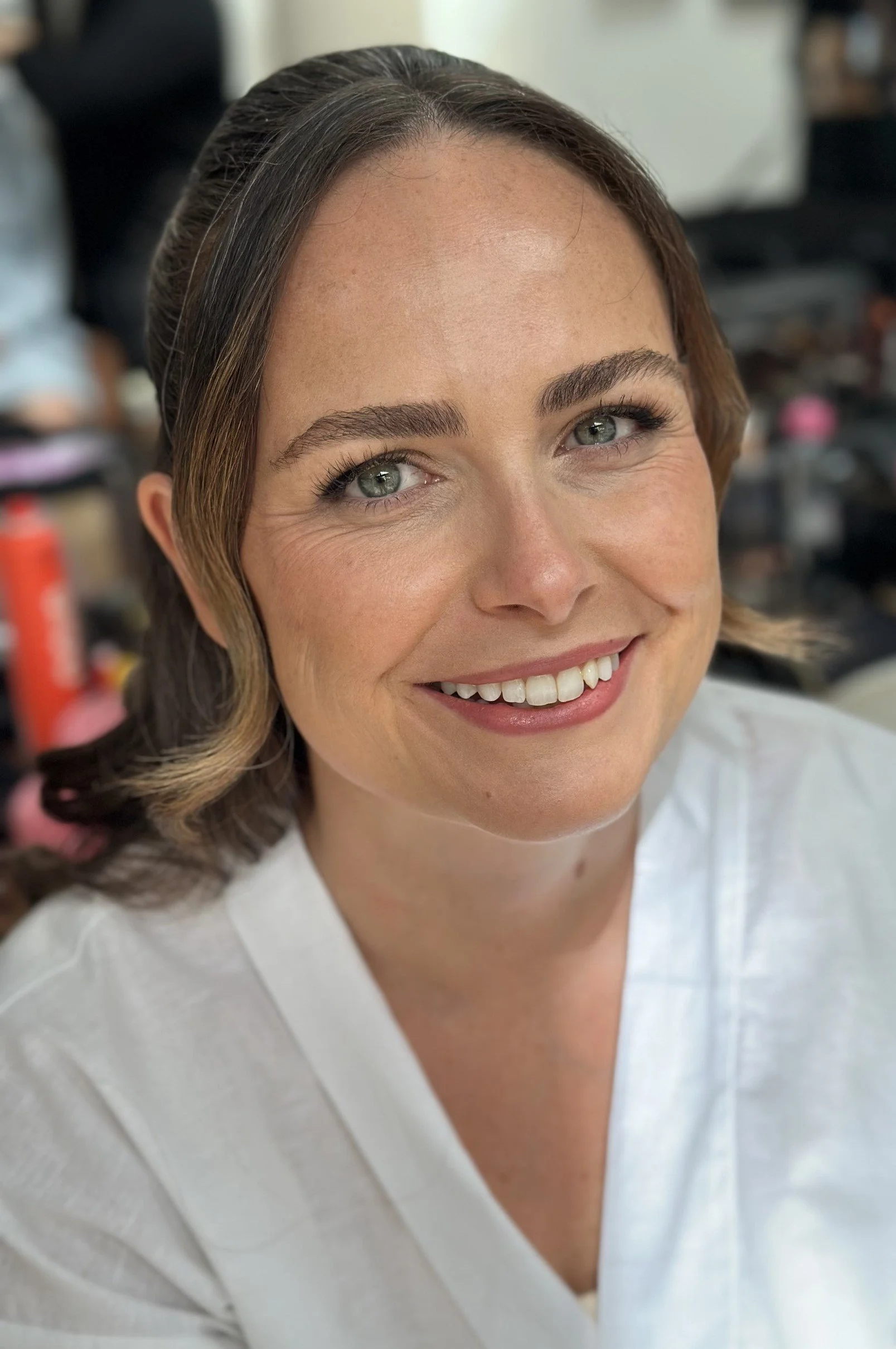 Close-up of a smiling woman with light skin, green eyes, and brown hair styled in loose waves, wearing a white top, in a room with various objects blurred in the background.