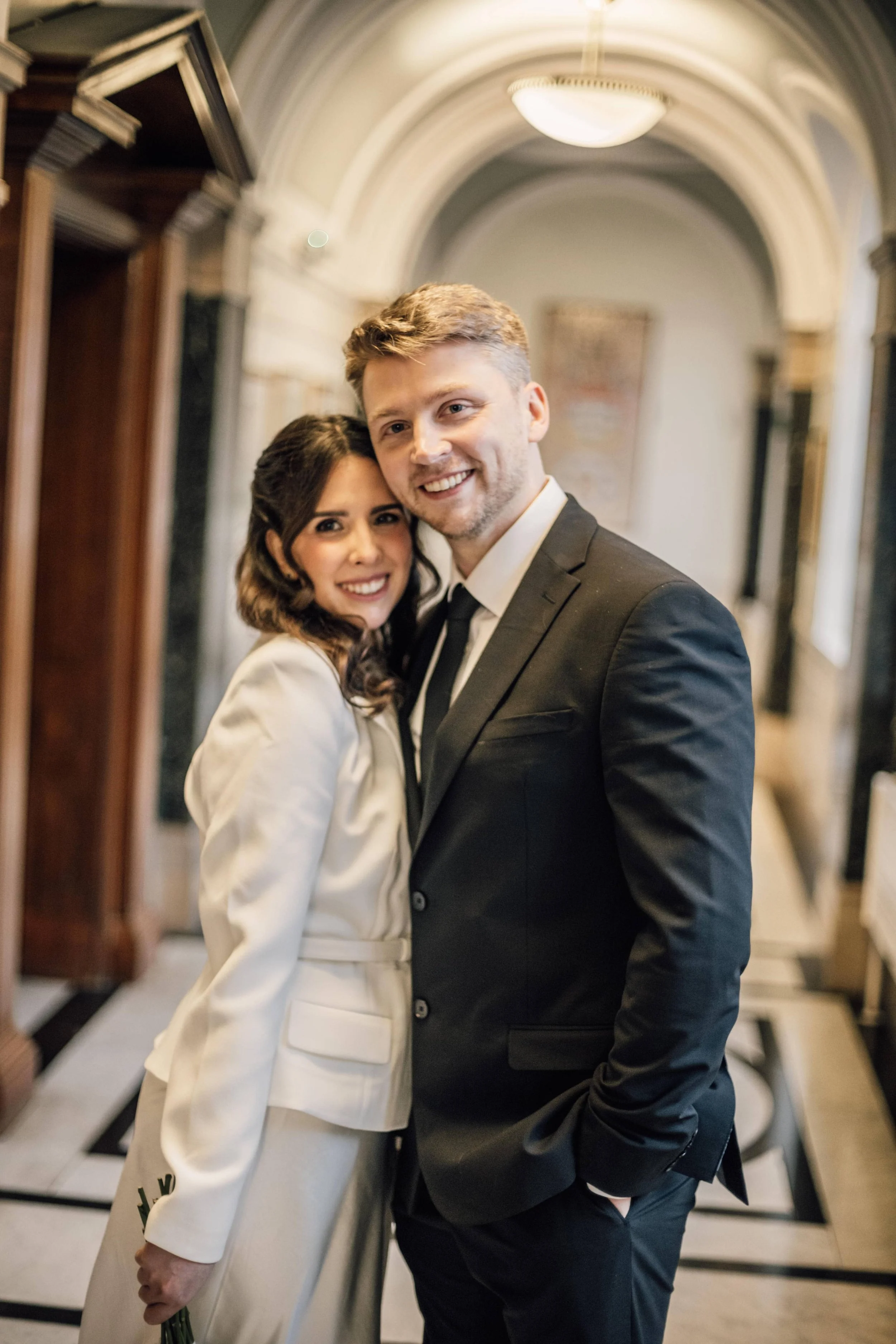 A smiling bride in a white wedding suit and a groom in a black suit standing close together inside a grand, arched hallway with ornate decor. They have just got married at Marylebone Town Hall in London.