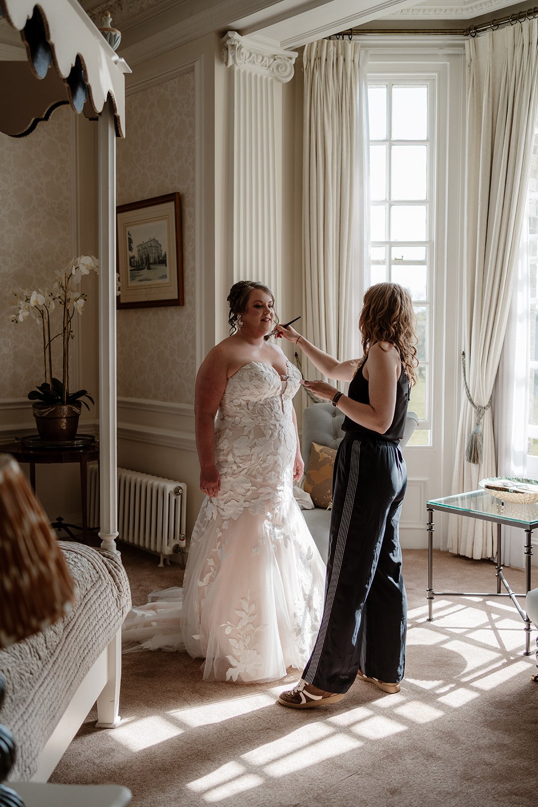 bride standing in a white wedding dress in front of a window, having her bridal makeup touch-ups done