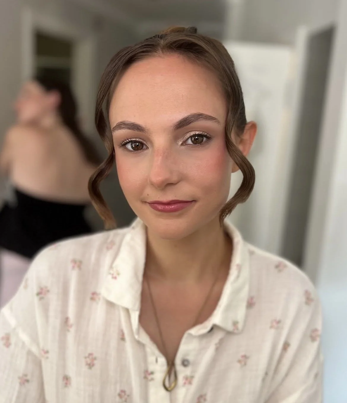 Close-up of a young woman with styled hair, soft glam makeup, and a floral blouse, smiling at the camera.