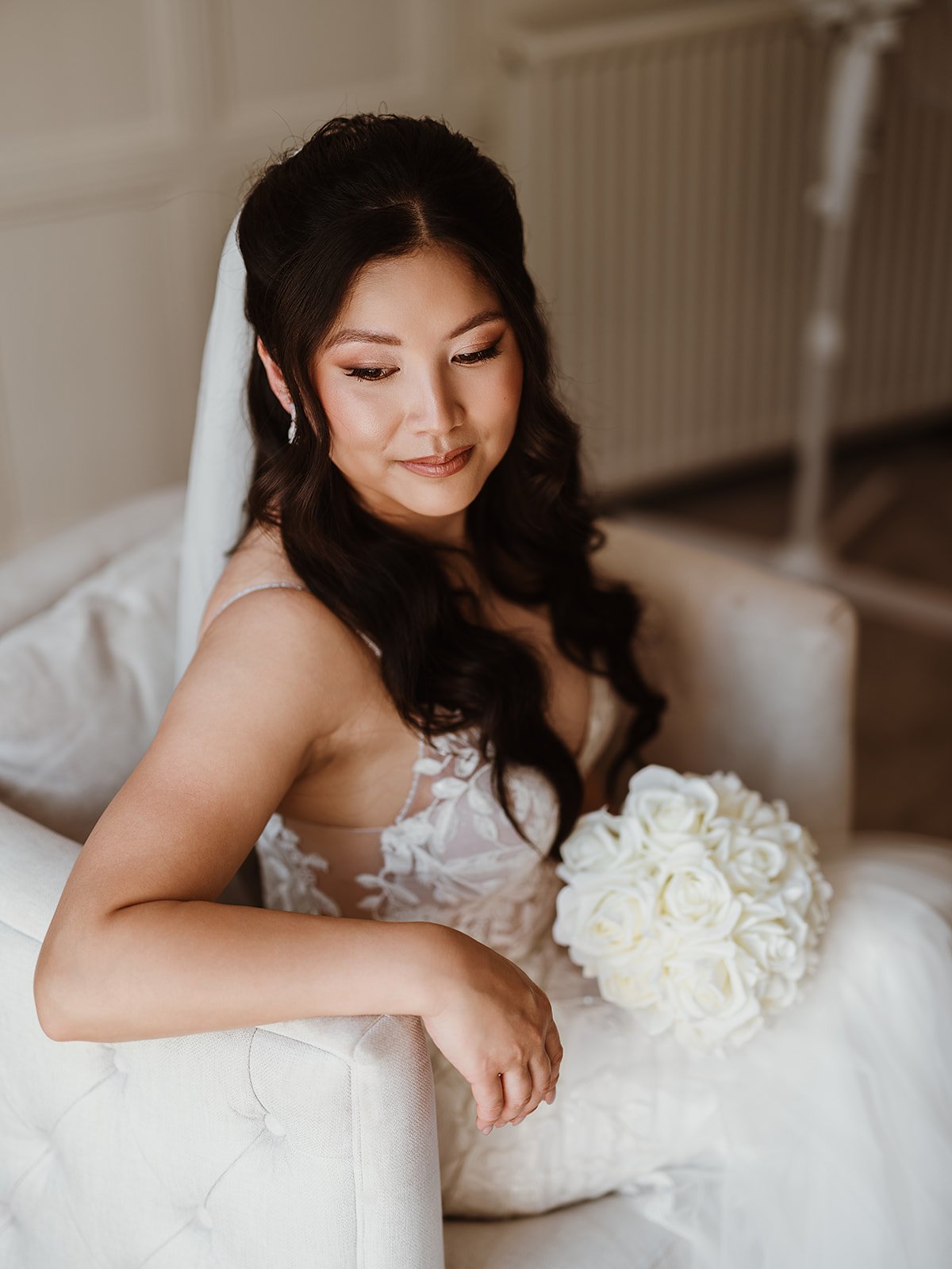a bride on her wedding day sits in a white armchair, wearing soft glam makeup and romantic, wavy hairstyle