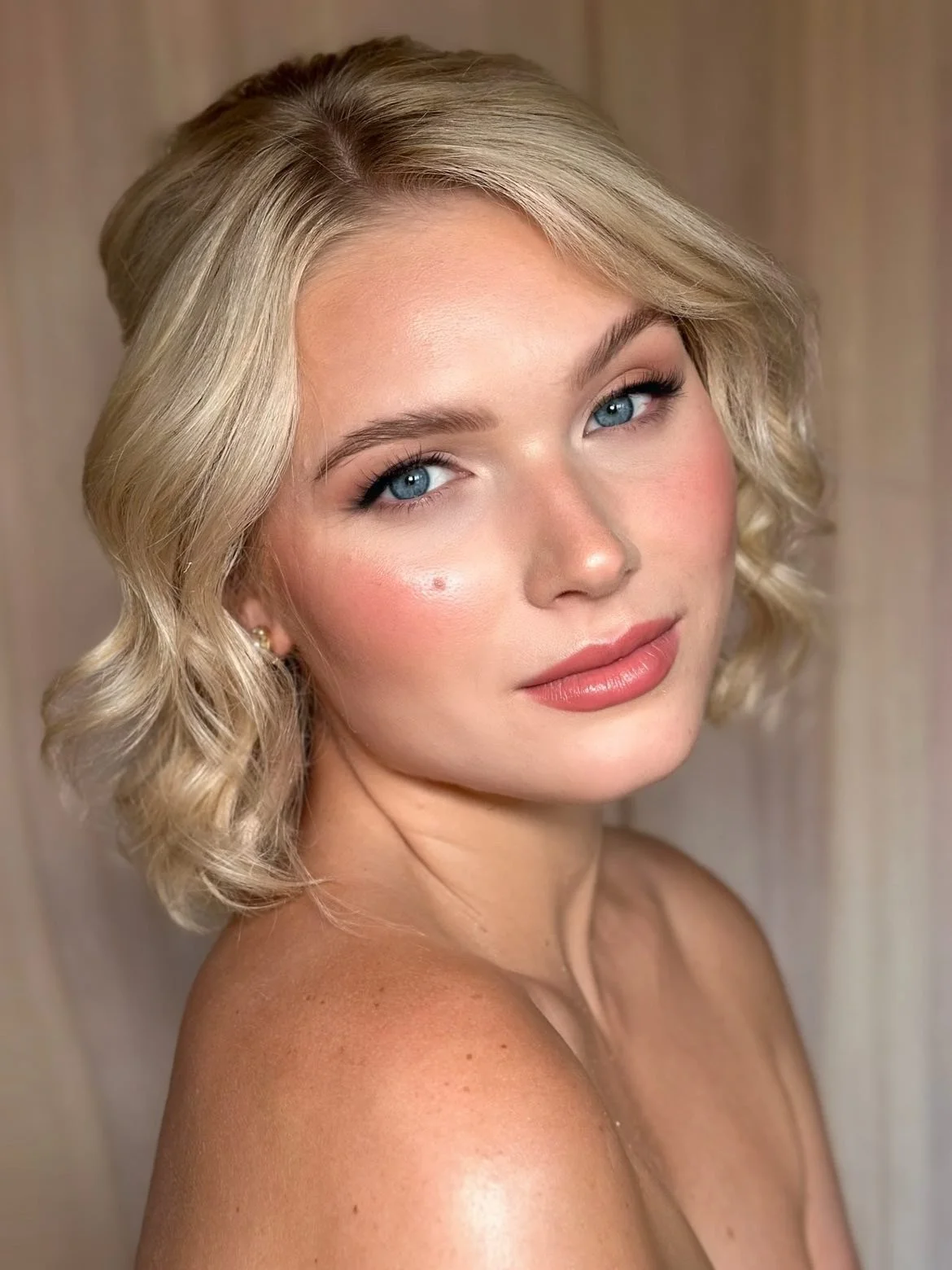 Close-up portrait of a blonde woman with wavy hair, blue eyes, natural makeup, and nude lipstick, looking at the camera.