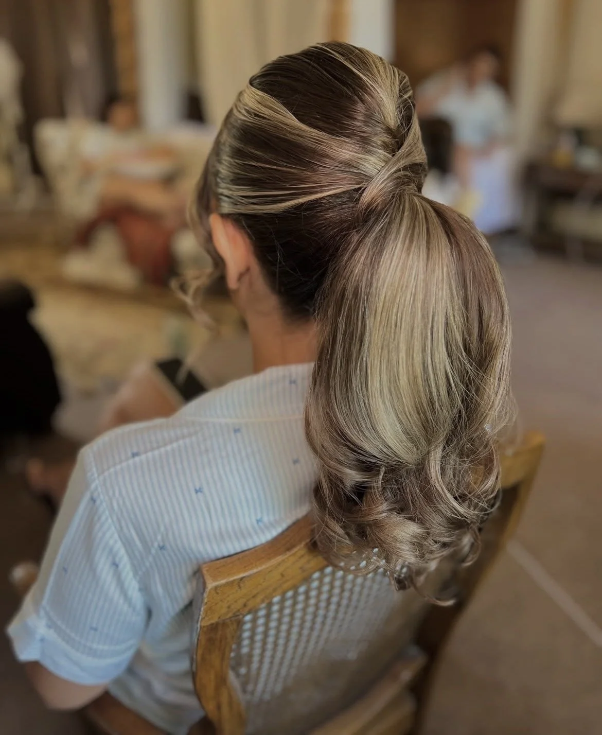 Back of a woman with blonde highlighted hairstyles in a ponytail with waves, sitting on a wooden chair indoors.