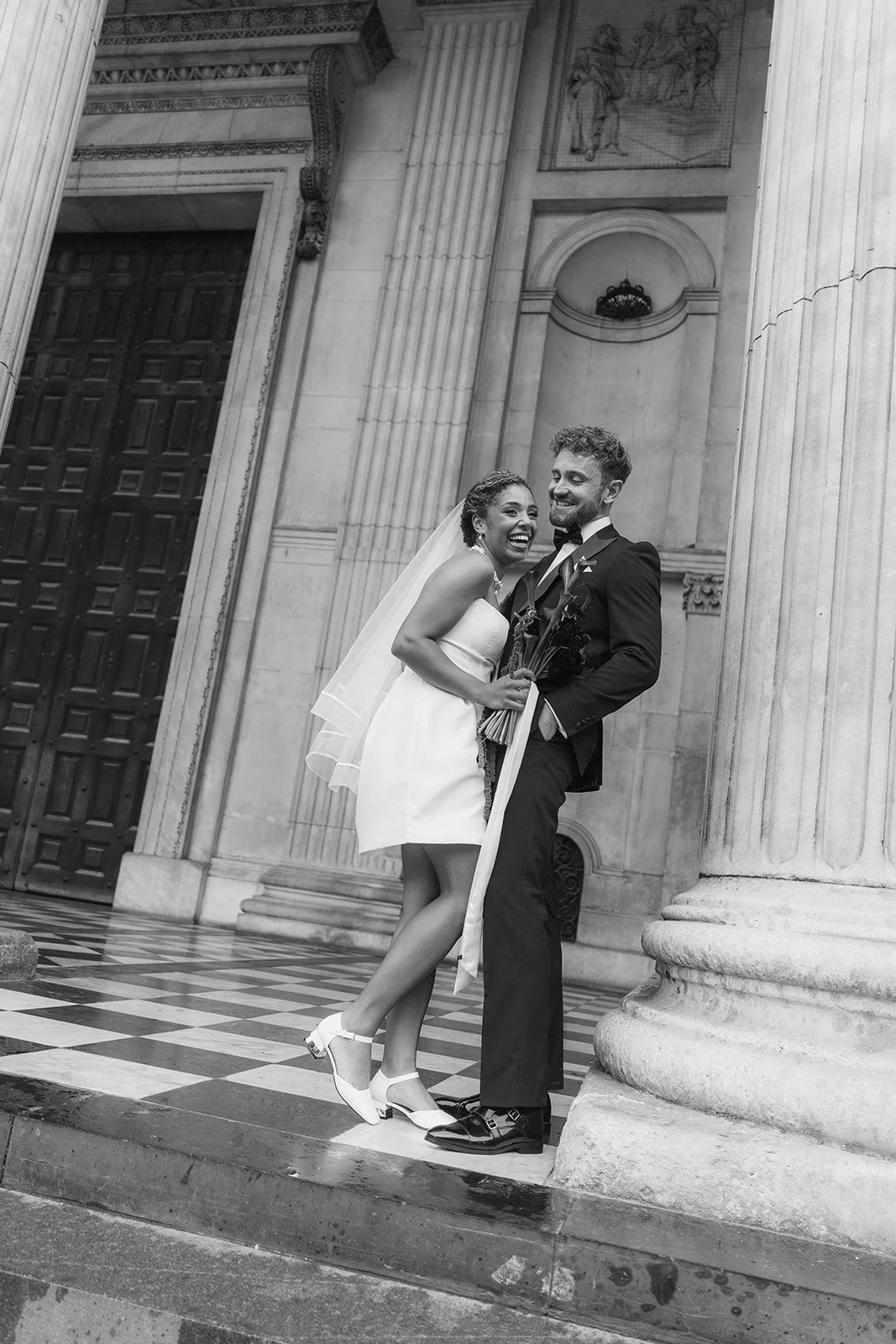 Black and white photo of a happy bride and groom standing together outside a St Paul's Cathedral, London, smiling and holding flowers.