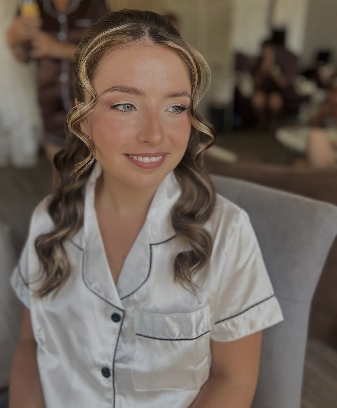 A bride with hair styled in soft waves, smiling and wearing satin pyjamas on a wedding morning.