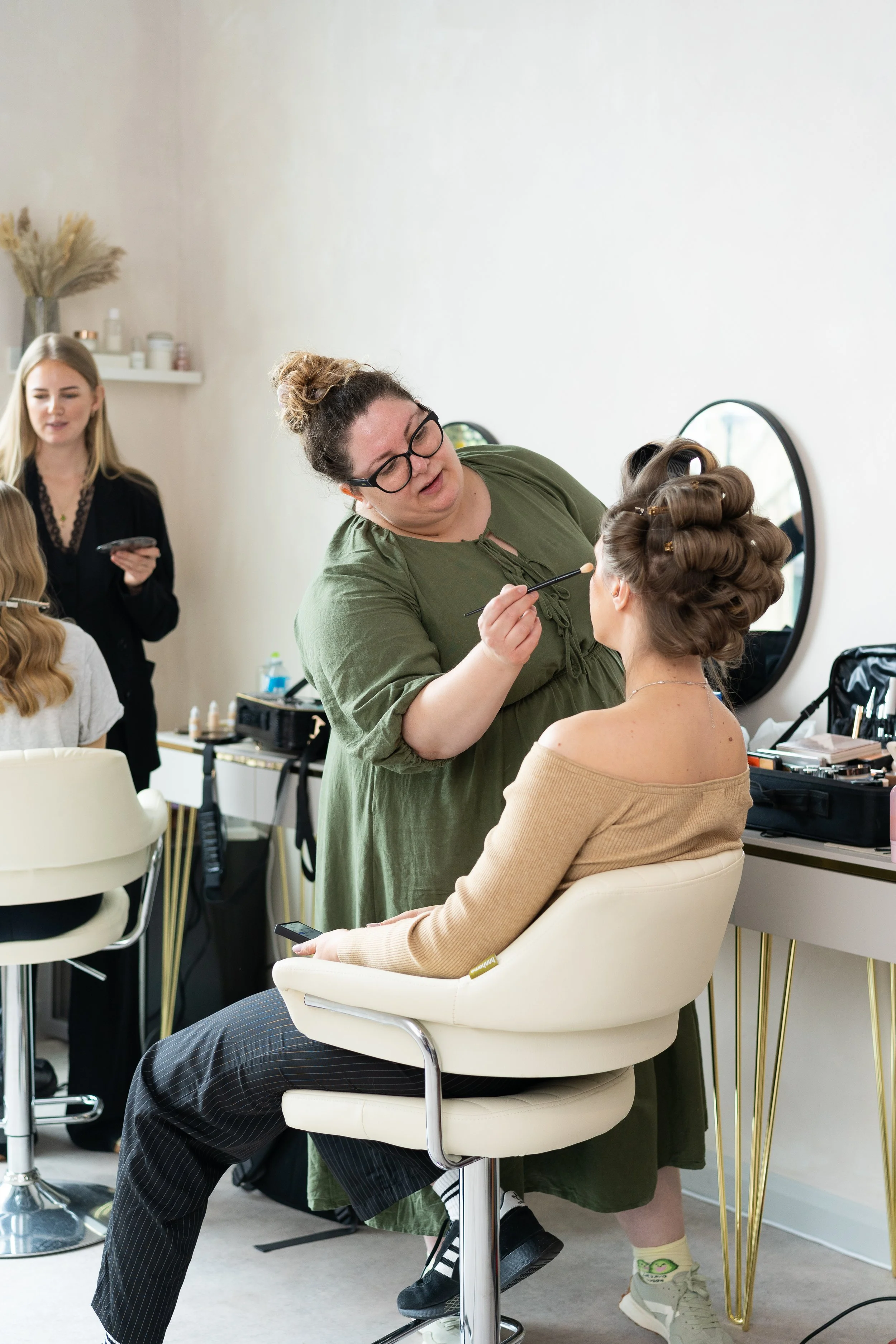 A makeup artist applies makeup to a woman seated in a makeup chair, with other people and makeup tools visible in the background.