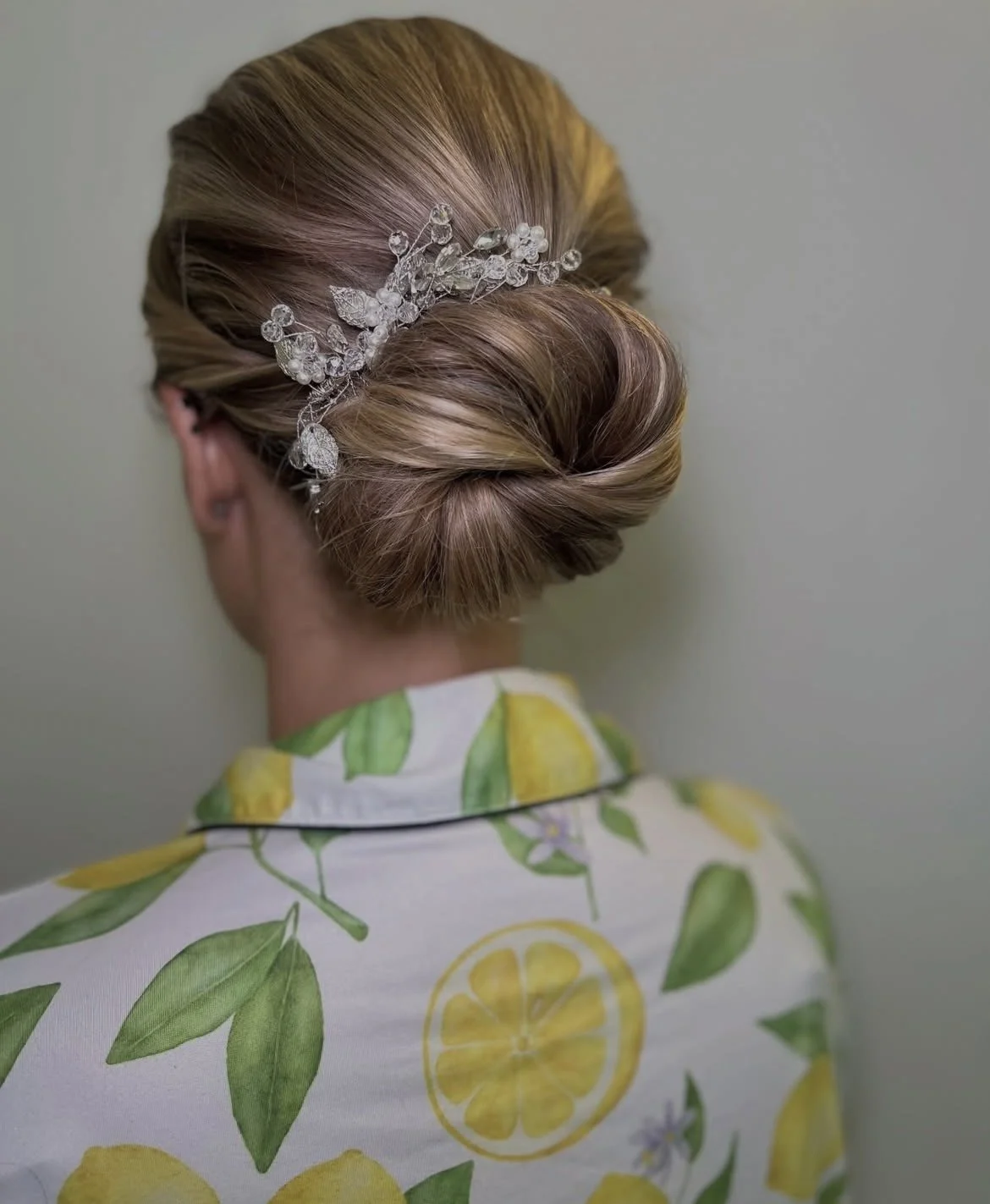Back view of a woman with blonde hair styled in an elegant updo with a decorative silver hairpiece, wearing a white shirt with a lemon and lemon leaf pattern.