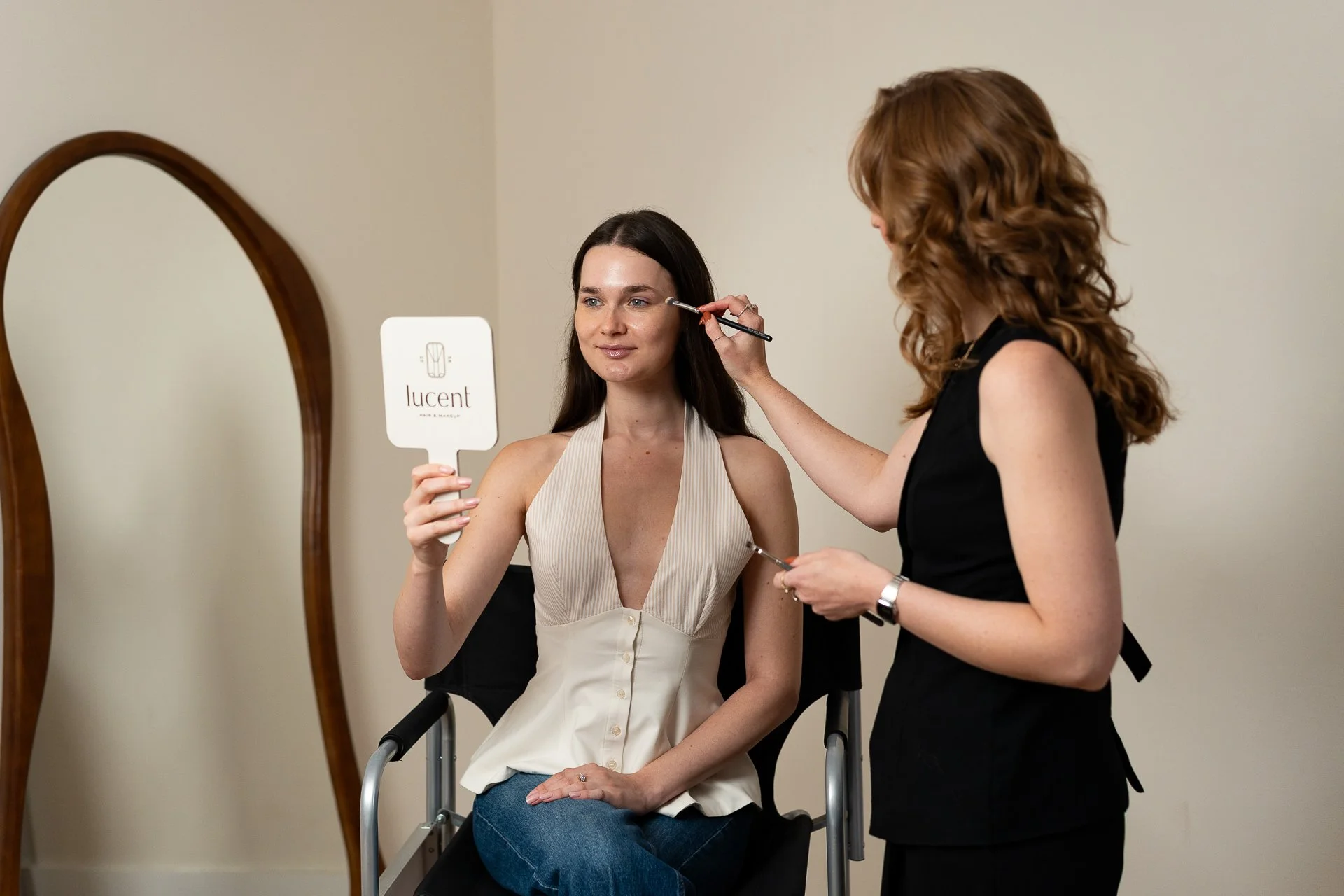 A makeup artist applies makeup to a woman as she looks at her reflection in a handheld mirror