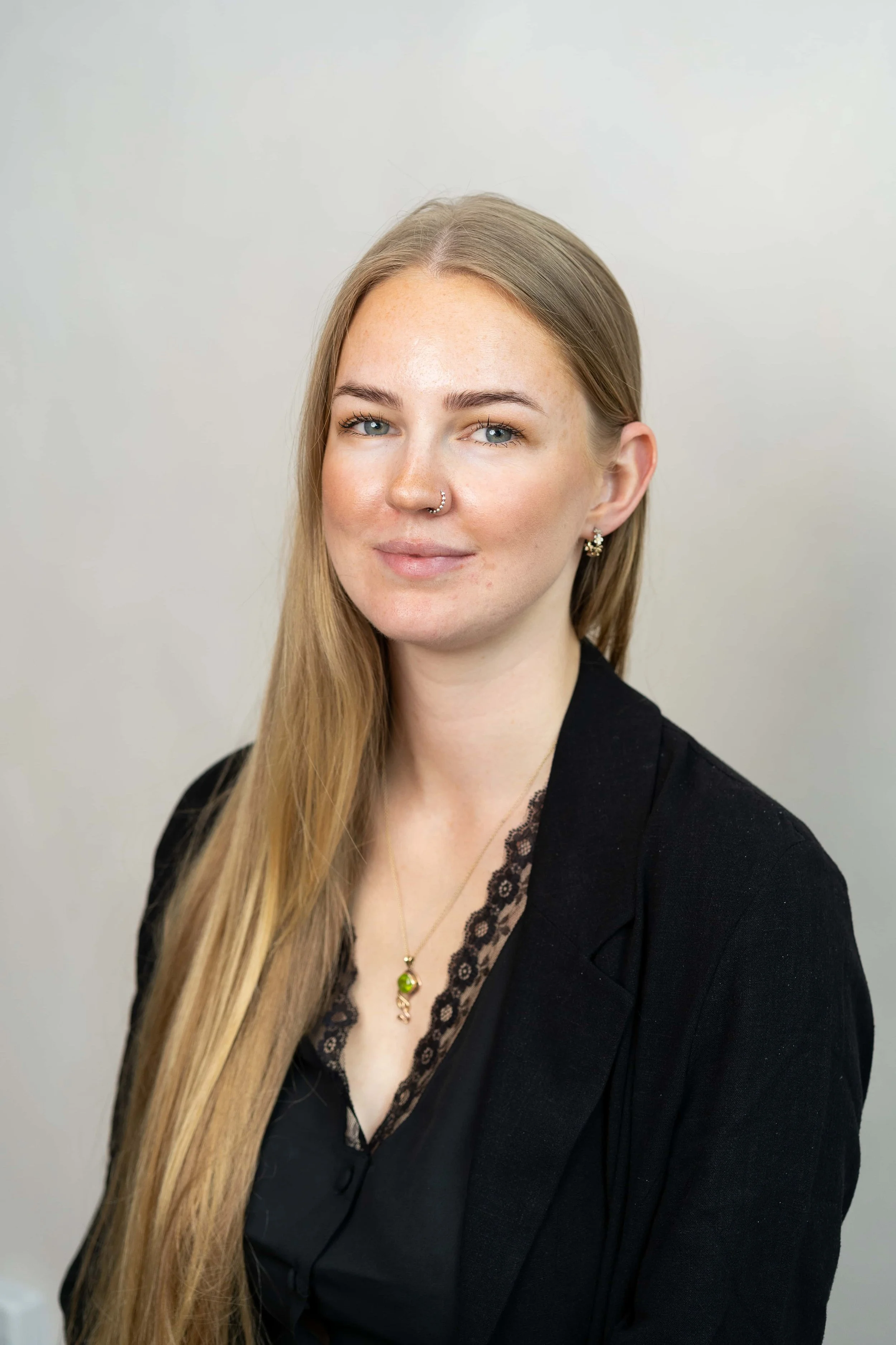 Portrait of a young woman with long blonde hair, blue eyes, wearing a black blazer, lace top, and jewellery, against a plain background.