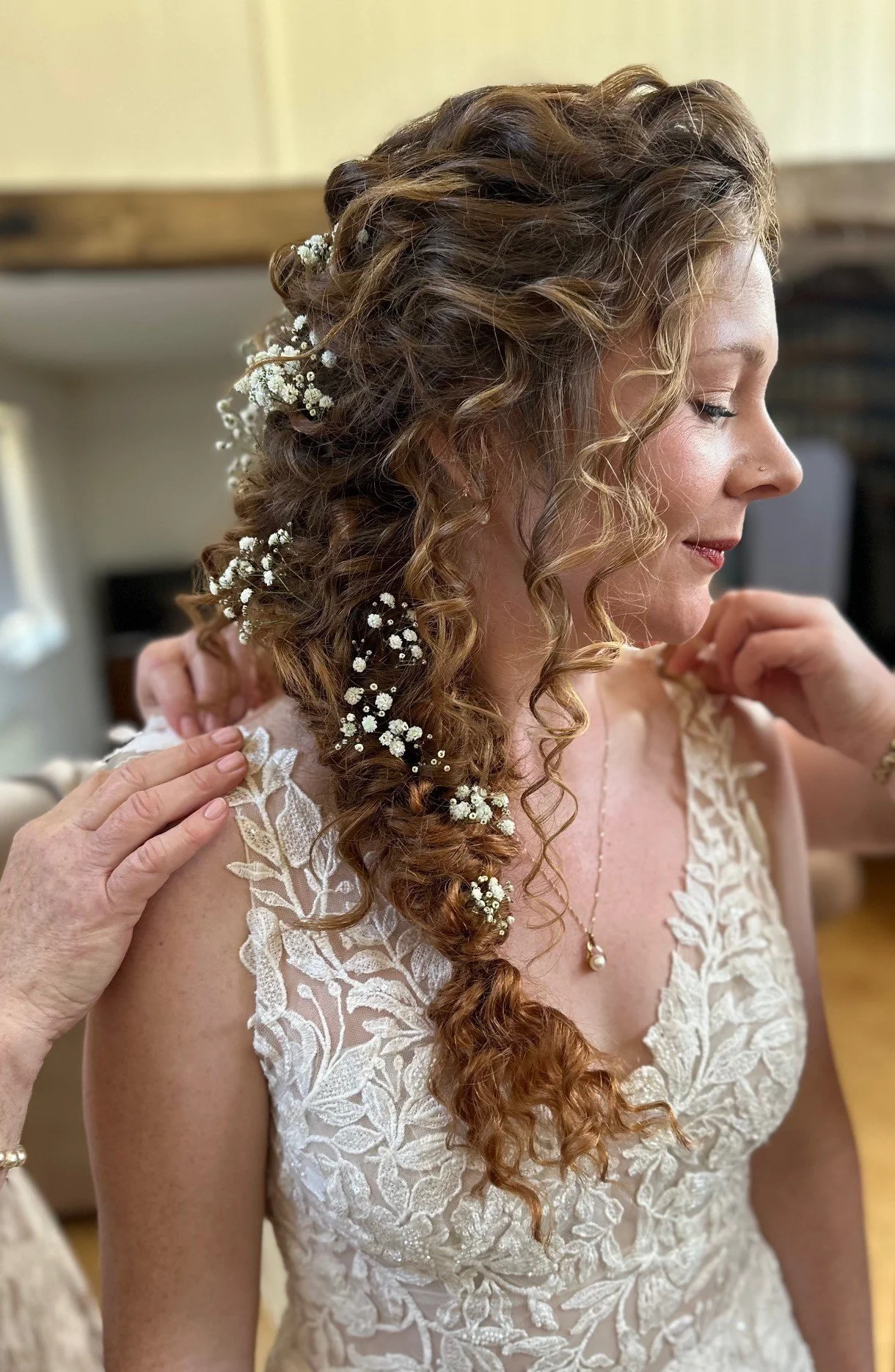 A bride with curly reddish-brown hair adorned with small white flowers during a wedding or special event, wearing a lace dress and a pearl necklace.