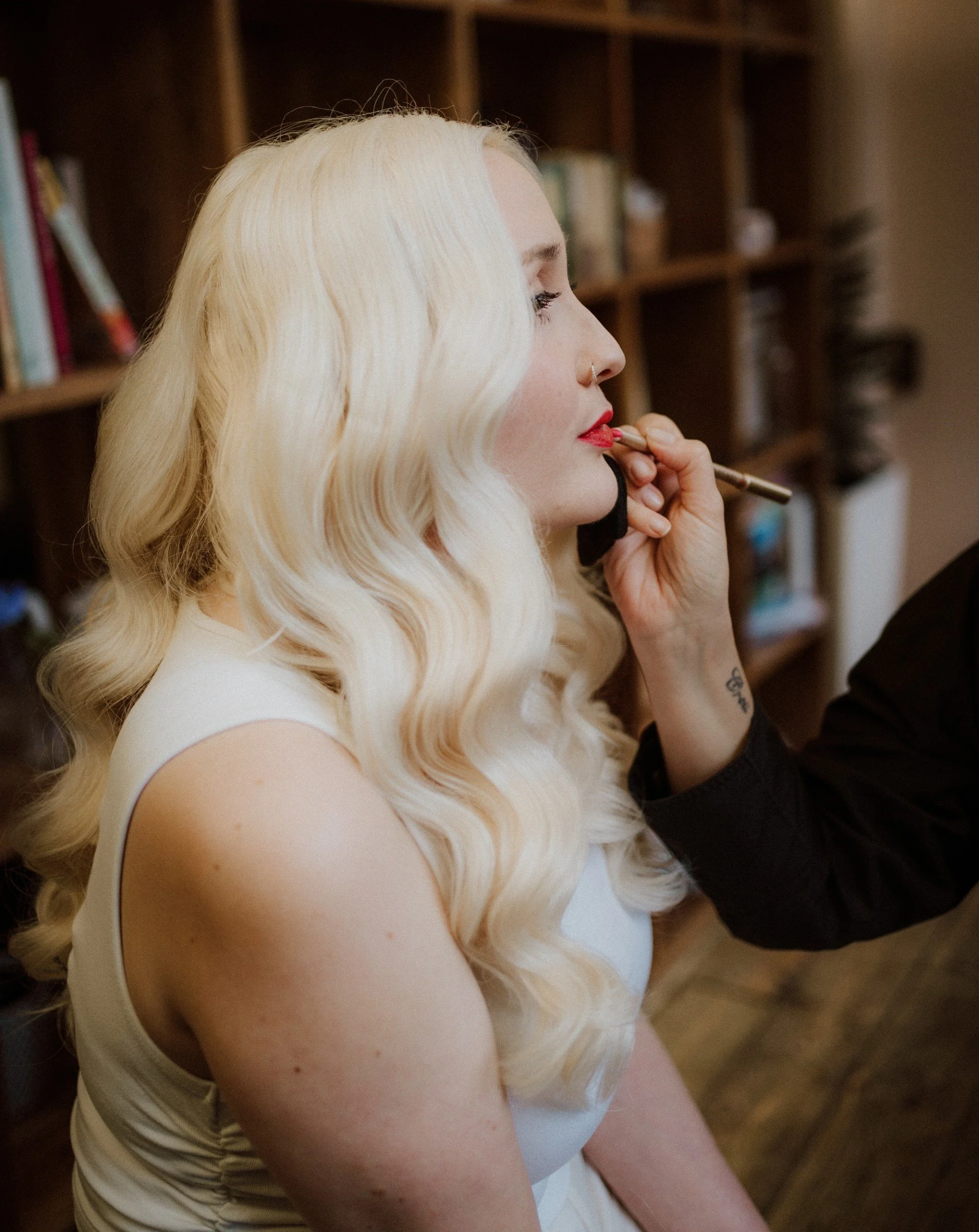 A woman with long, wavy platinum blonde hair styled in Hollywood waves, getting her makeup done, with a makeup artist applying red lipstick on her lips and holding a makeup brush, in front of a wooden bookshelf filled with books.
