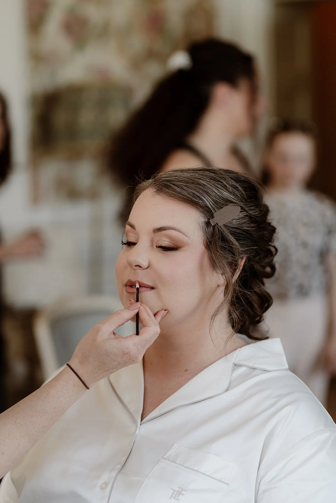 a bride has her wedding day lipstick applied as she dits in a makeup chair, wearing white pyjamas