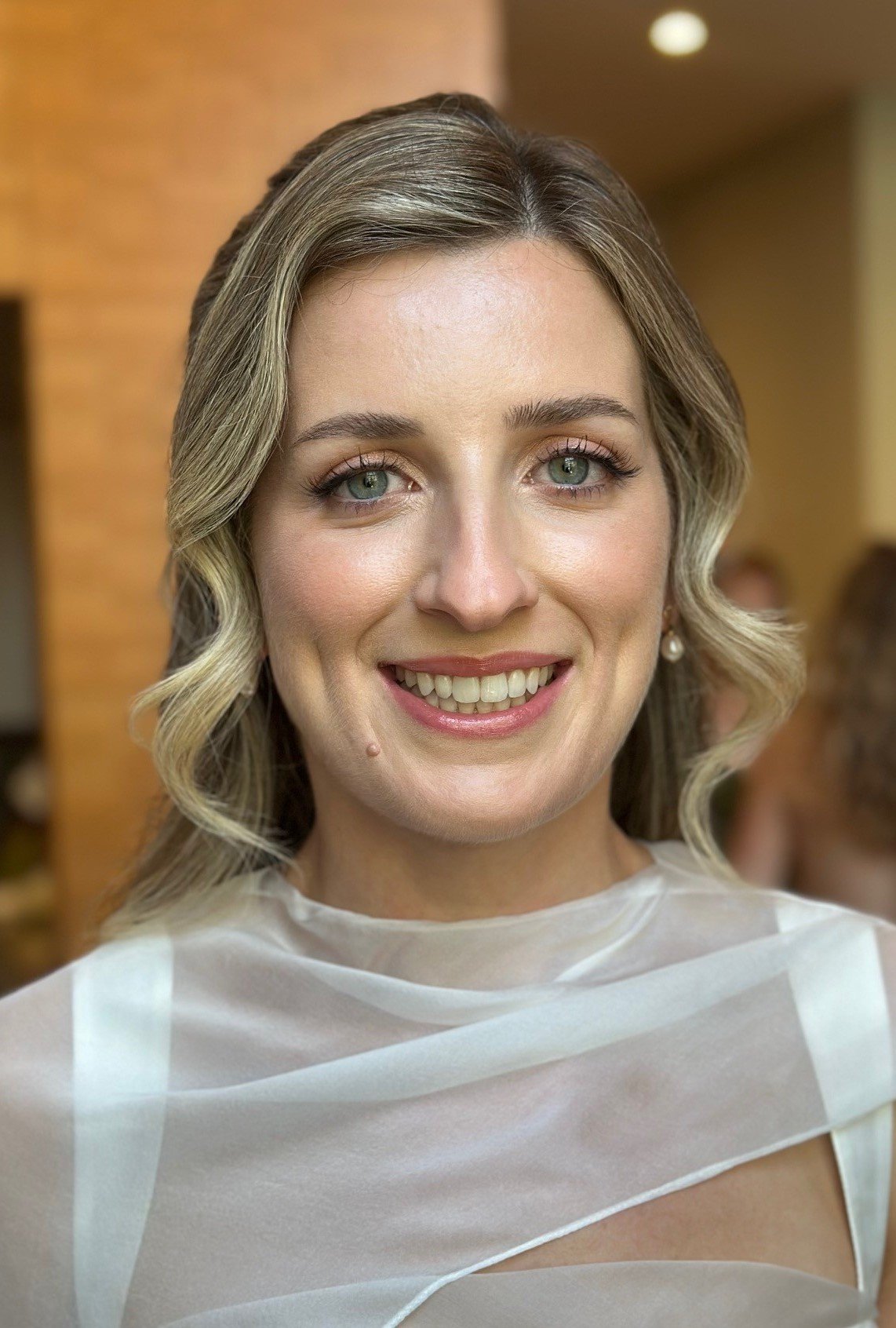 Close-up of a bride with shoulder-length wavy blonde hair and green eyes, smiling, wearing pearl earrings and a sheer white top, indoors.