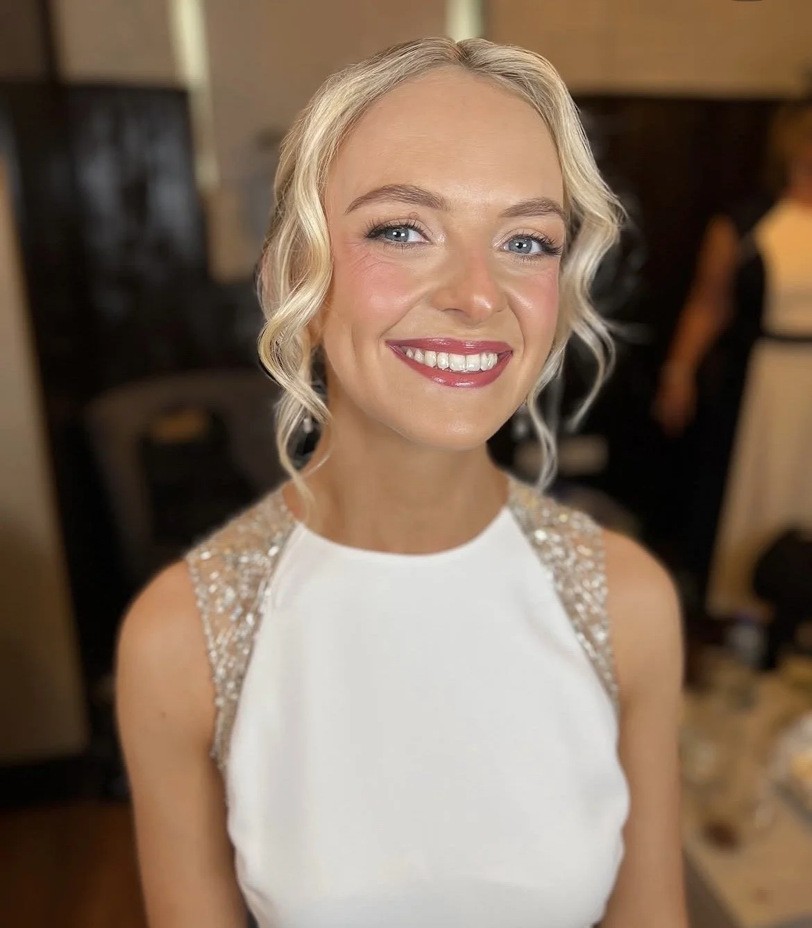 Close-up of a smiling bride with blonde hair styled in an undo with loose curls, wearing a white wedding dress with beaded details on the shoulders, in an indoor setting.